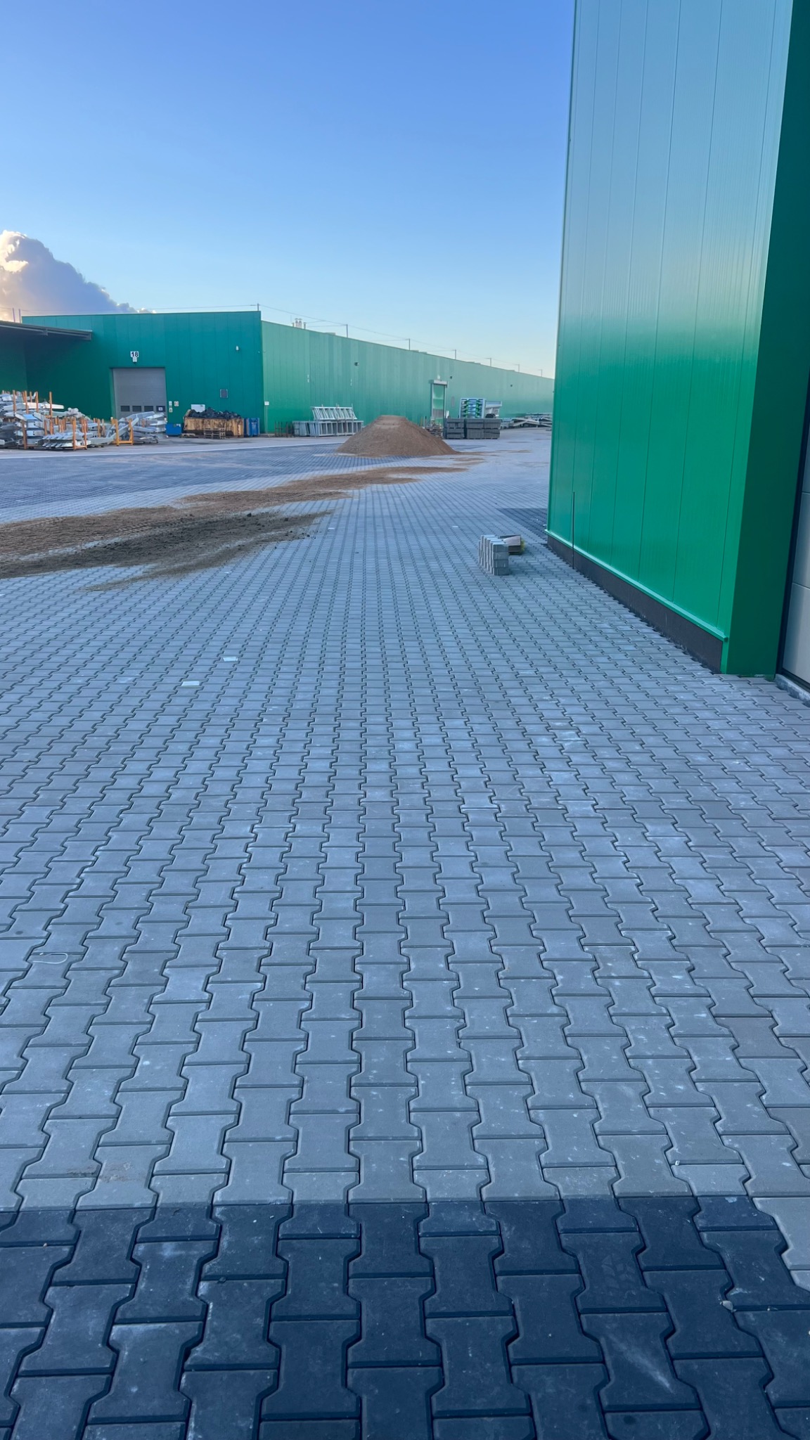 Szary chodnik z kostki brukowej typu 'I' prowadzący do zielonego budynku przemysłowego, widoczny niewielki stos piasku i palety w tle.