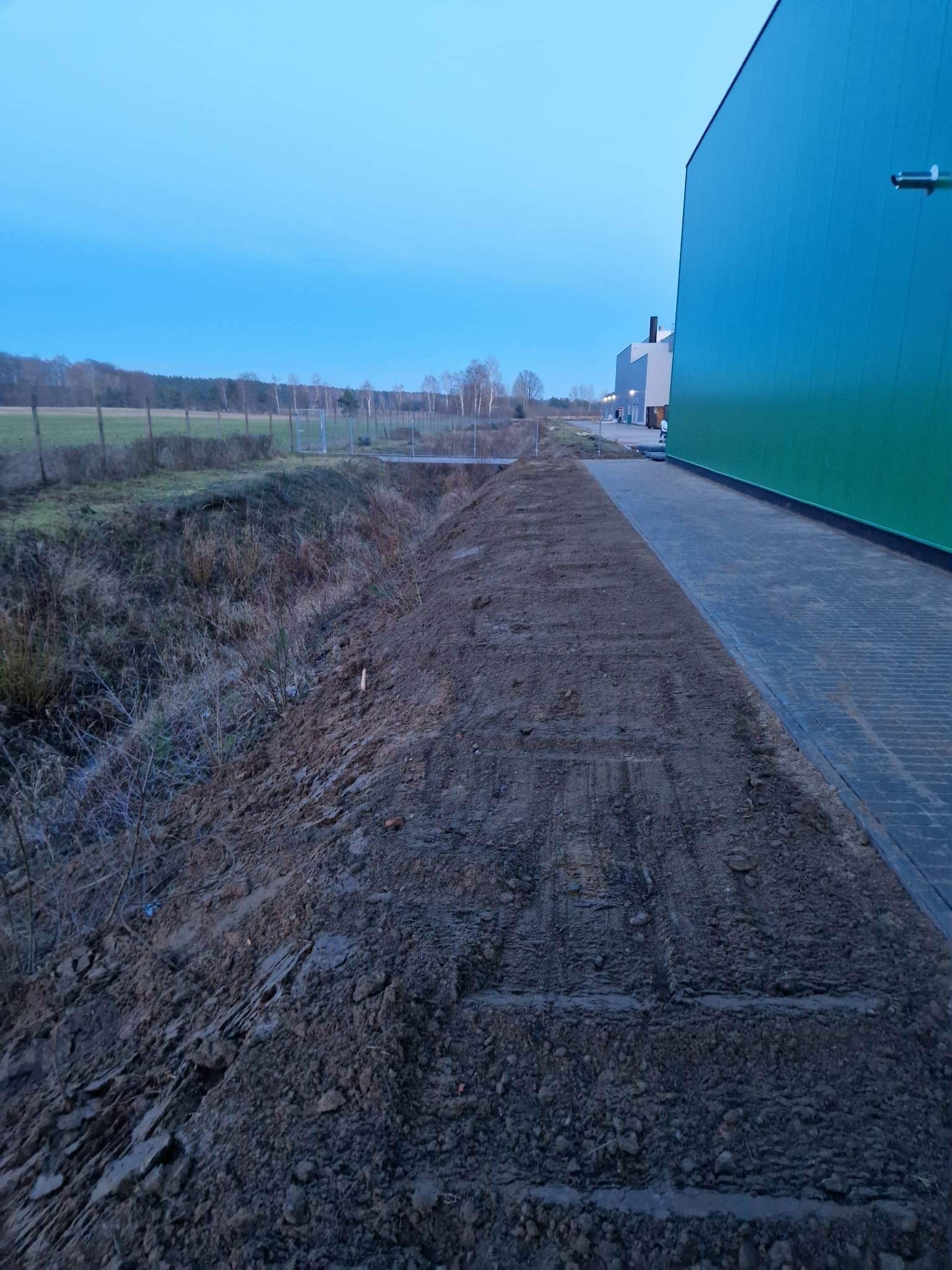 Przygotowany teren pod ścieżkę z kostki brukowej przy zielonej hali magazynowej, widoczne ślady po maszynach budowlanych. Tło stanowi krajobraz z drzewami i błękitne niebo.
