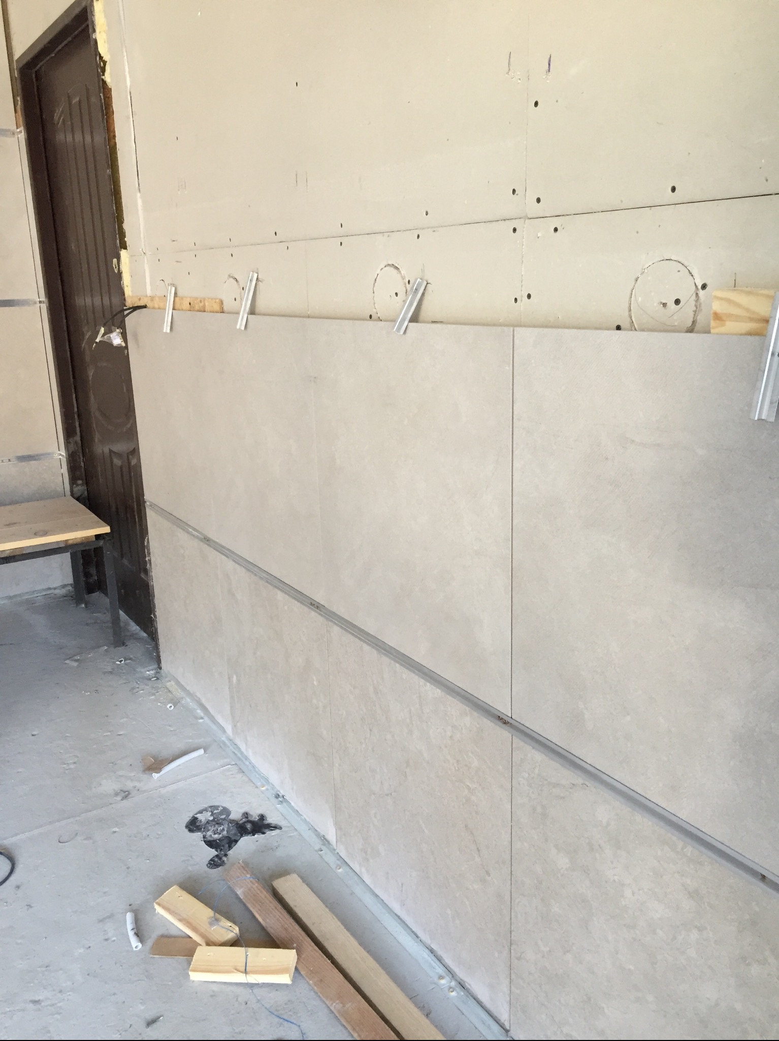 Montaż płytek ceramicznych na ścianie z płyt kartonowo-gipsowych, widoczne profile montażowe i drewniane kliny, surowy stan podłogi z resztkami zaprawy.