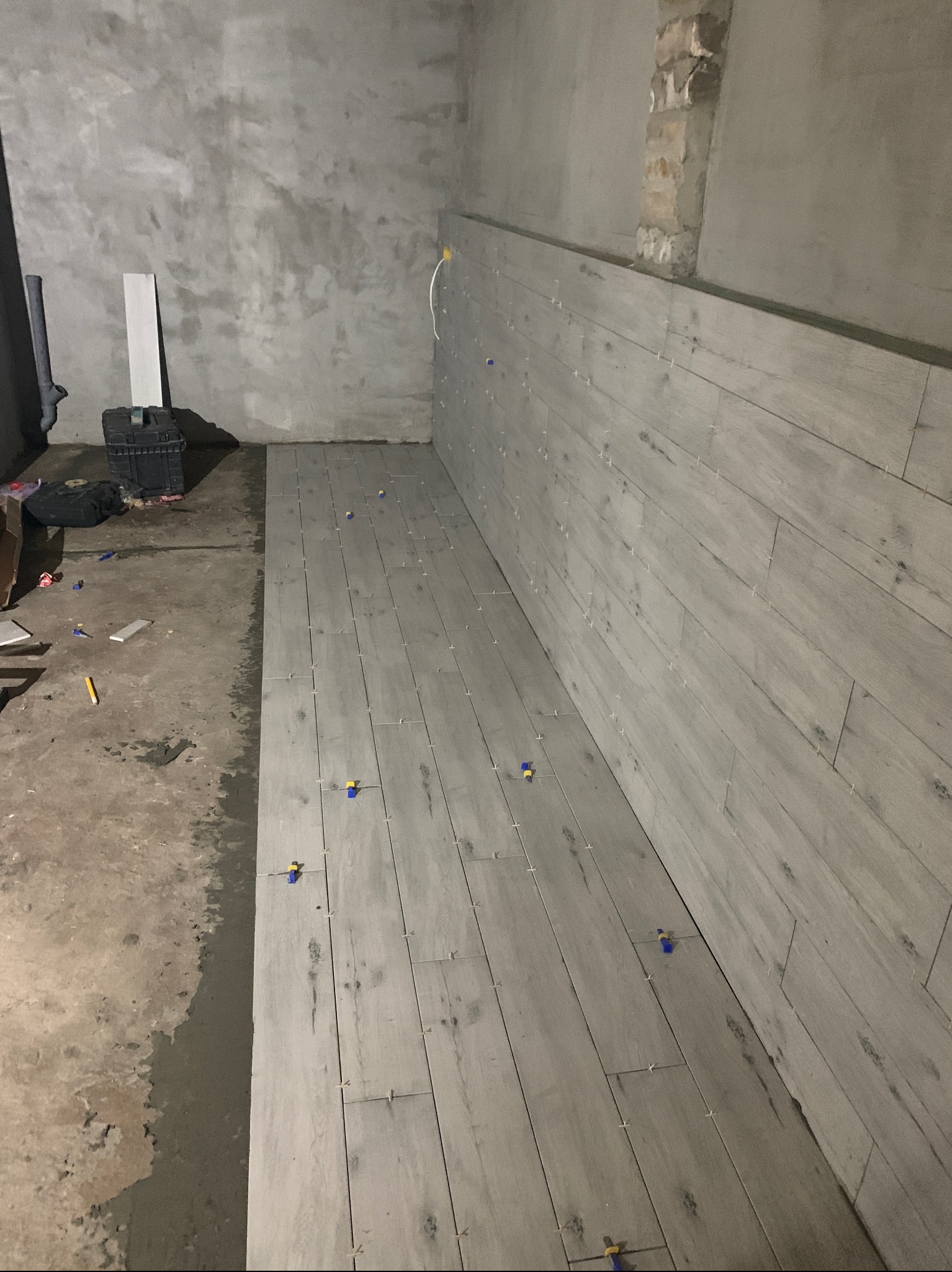Układanie szarych płytek imitujących drewno na podłodze i ścianie w surowym pomieszczeniu, widoczne krzyżyki dystansowe i kliny poziomujące, w tle niezabezpieczona ściana z cegły.
