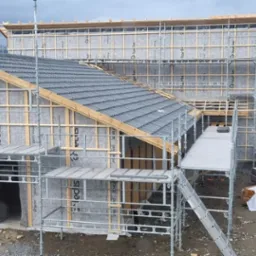 Wielopoziomowe rusztowanie otacza budynek w trakcie wznoszenia, z widoczną konstrukcją drewnianą, szarą membraną izolacyjną i częściowo położonym ciemnoszarym dachem. W tle jezioro i góry.