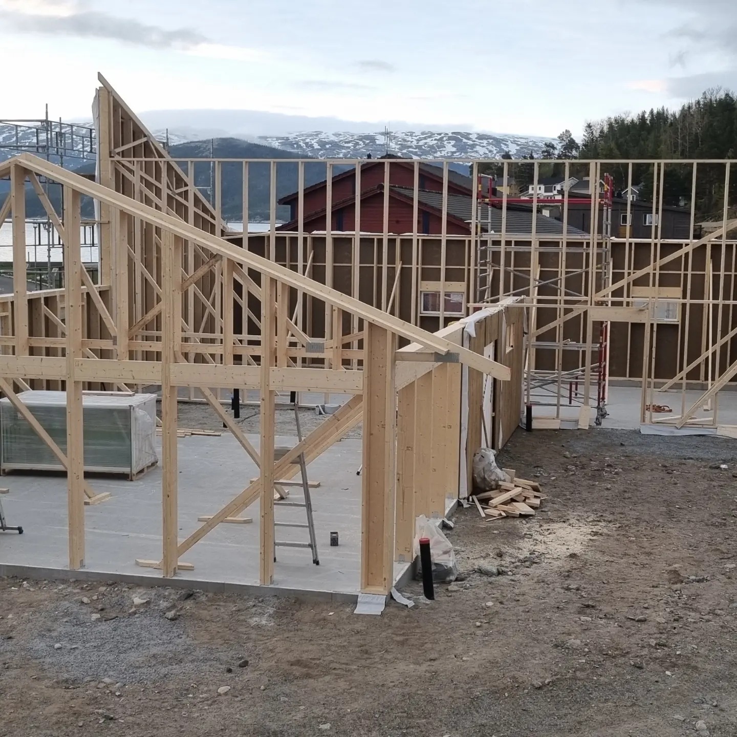 Wznoszenie drewnianej konstrukcji domu w trakcie budowy, widoczne elementy szkieletu, w tle góry i zabudowa mieszkalna.