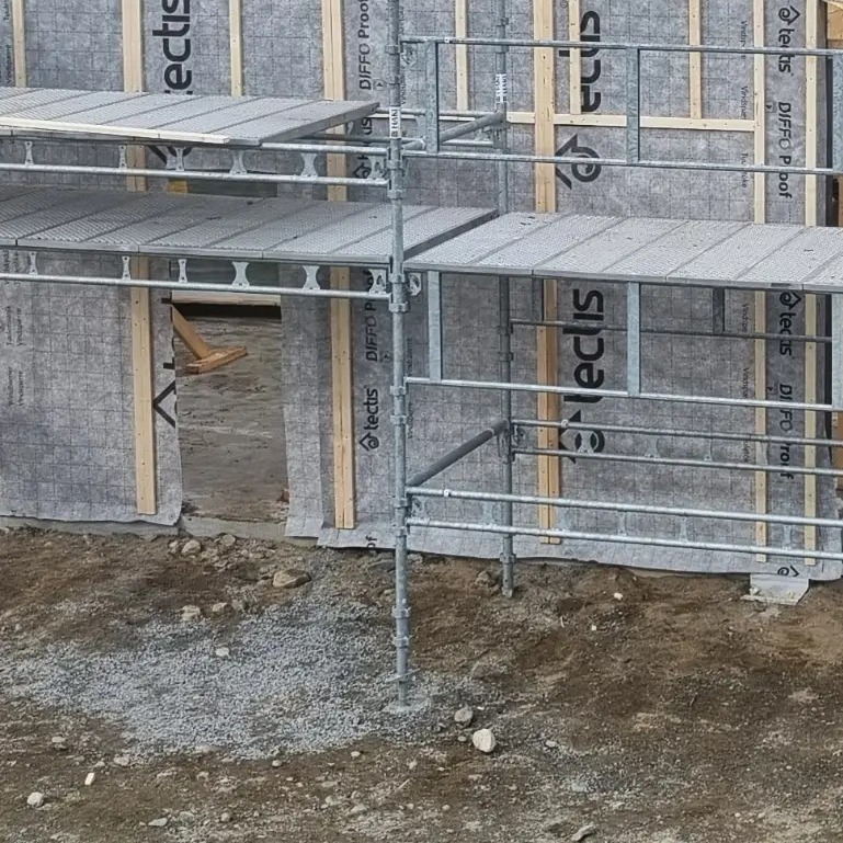Wznoszona konstrukcja budynku z widocznymi rusztowaniami, drewnianymi elementami szkieletu i szarą membraną izolacyjną na zewnątrz, z widocznym fragmentem terenu budowy z gruzem i ziemią.