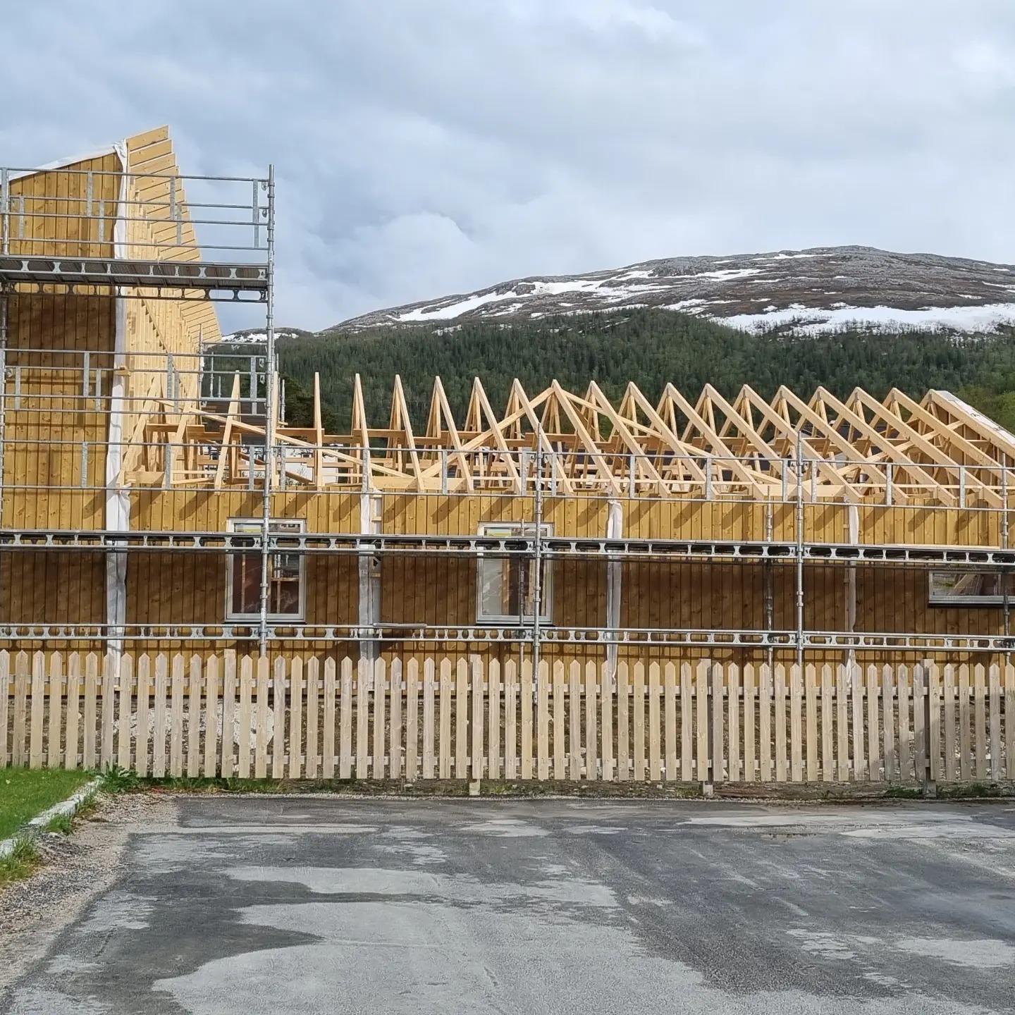 Wznoszenie konstrukcji szkieletowej domu z widocznym drewnianym szkieletem dachu, otoczonego rusztowaniem, z drewnianym płotem w tle i górą z resztkami śniegu na horyzoncie.