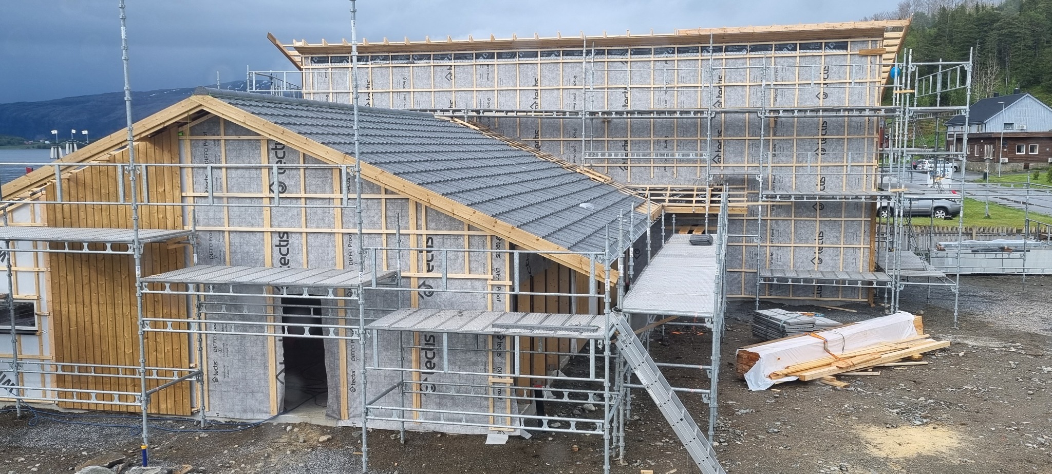 Wielopoziomowe rusztowanie otacza budynek w trakcie wznoszenia, z widoczną konstrukcją drewnianą, szarą membraną izolacyjną i częściowo położonym ciemnoszarym dachem. W tle jezioro i góry.