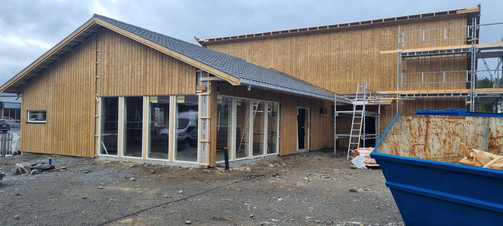 Budowa domu z drewnianą elewacją w trakcie realizacji, widoczne rusztowania i kontener na odpady budowlane, okna z białymi ramami wstawione w surową konstrukcję.