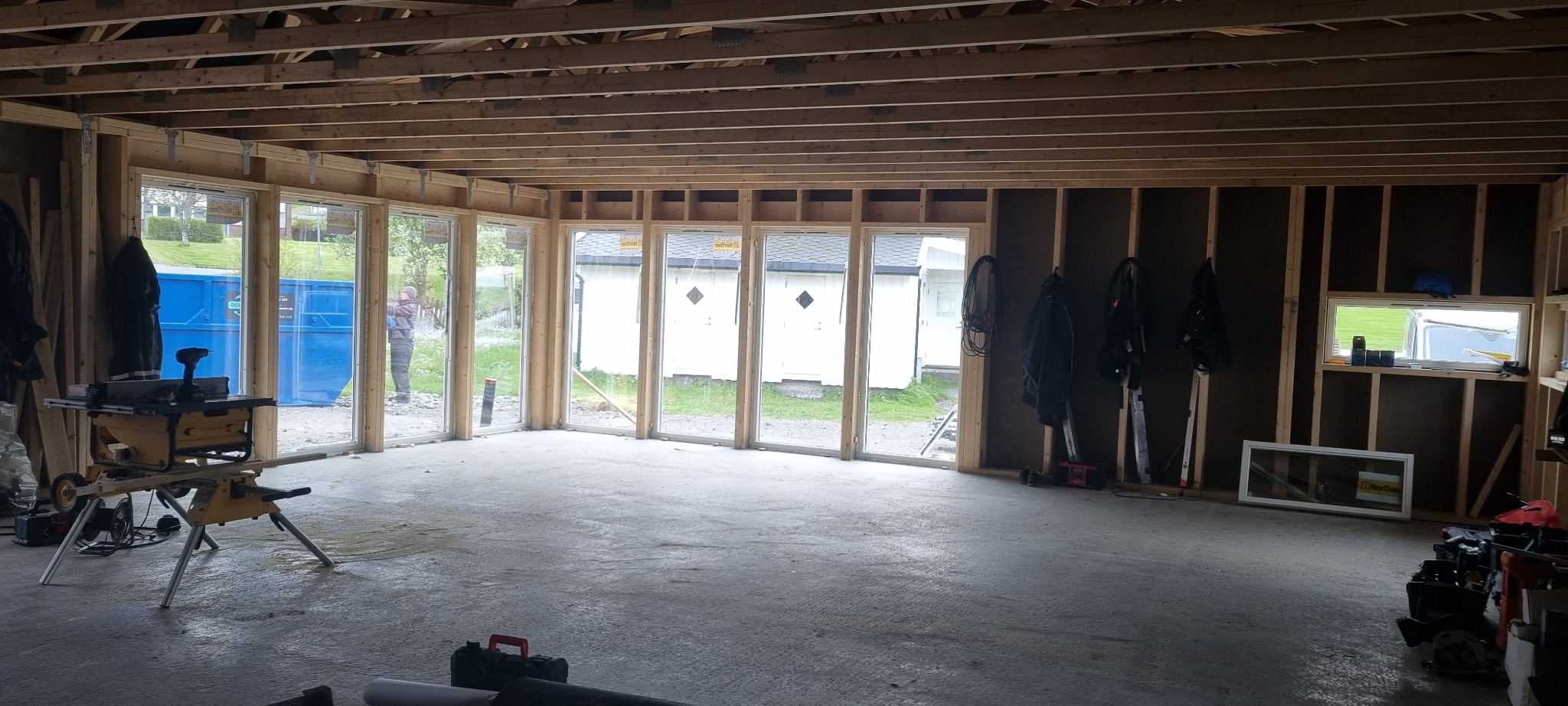 Wnętrze w stanie surowym otwartym, widoczne drewniane ramy konstrukcyjne ścian i stropu, duże okna z widokiem na zewnątrz, narzędzia budowlane i niebieski kontener na śmieci.