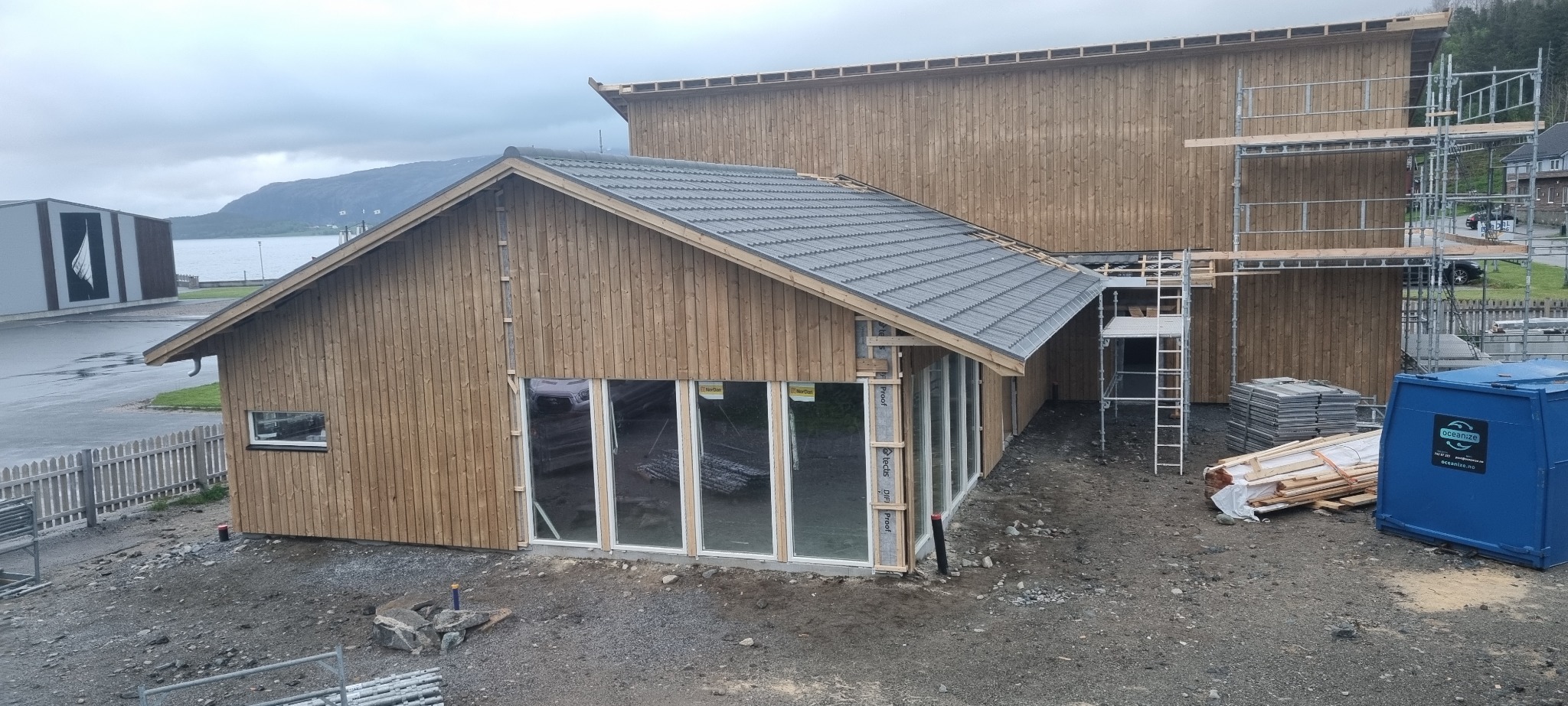 Budynek w trakcie budowy z drewnianą elewacją, szarą dachówką i dużymi oknami, widoczne rusztowania i materiały budowlane w otoczeniu.