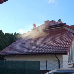 F. U. Alpina Mycie Malowanie Dachów. - Pracownik w pomarańczowym kombinezonie czyści dach z czerwonej dachówki pod ciśnieniem, widoczna para wodna, w tle zielone tuje i fragment zaparkowanego szarego samochodu.