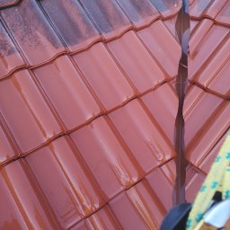 F. U. Alpina Mycie Malowanie Dachów. - Czerwona dachówka ceramiczna po umyciu, z widocznymi śladami wody i różnicą w kolorze między czystą a zabrudzoną powierzchnią, fragment rynny i liny zabezpieczającej w kadrze.
