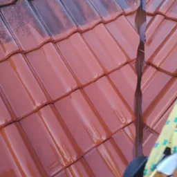 Czerwona dachówka ceramiczna po umyciu, z widocznymi śladami wody i różnicą w kolorze między czystą a zabrudzoną powierzchnią, fragment rynny i liny zabezpieczającej w kadrze.