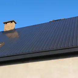 Ciemnoszary dach z blachy trapezowej, ceglany komin z daszkiem, fragment otynkowanej ściany budynku i fragment rusztowania na tle błękitnego nieba.