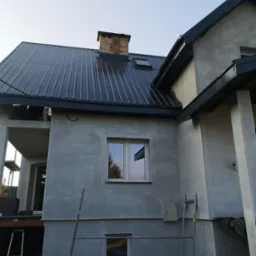 Dom w trakcie budowy lub remontu, z nowym, błyszczącym, ciemnym dachem z blachy trapezowej, widoczne rusztowania i surowa elewacja w kolorze szarym.