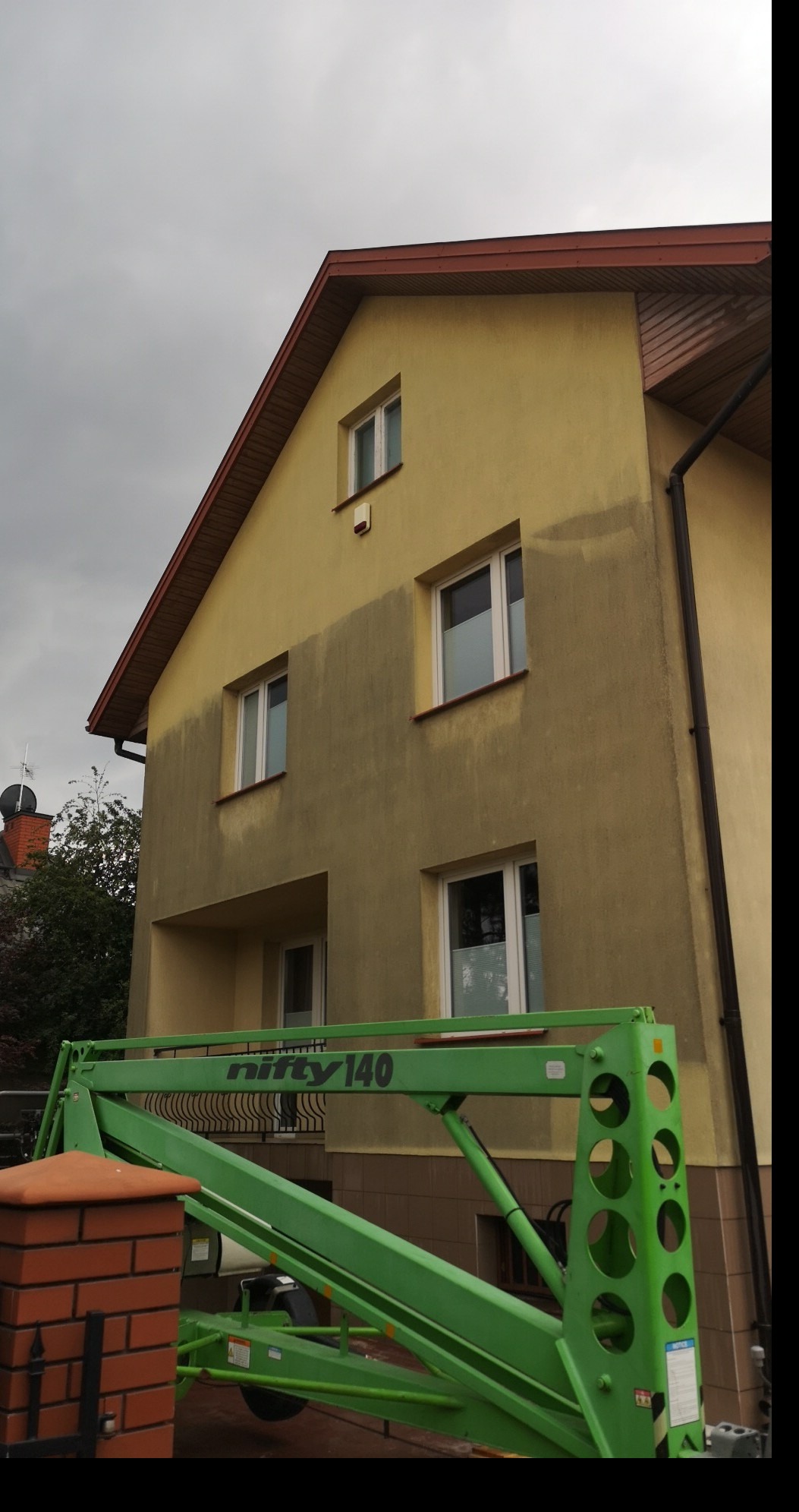 Dwukondygnacyjny dom z żółtą elewacją, częściowo umytą, widoczny z poziomu podnośnika koszowego Nifty 140.
