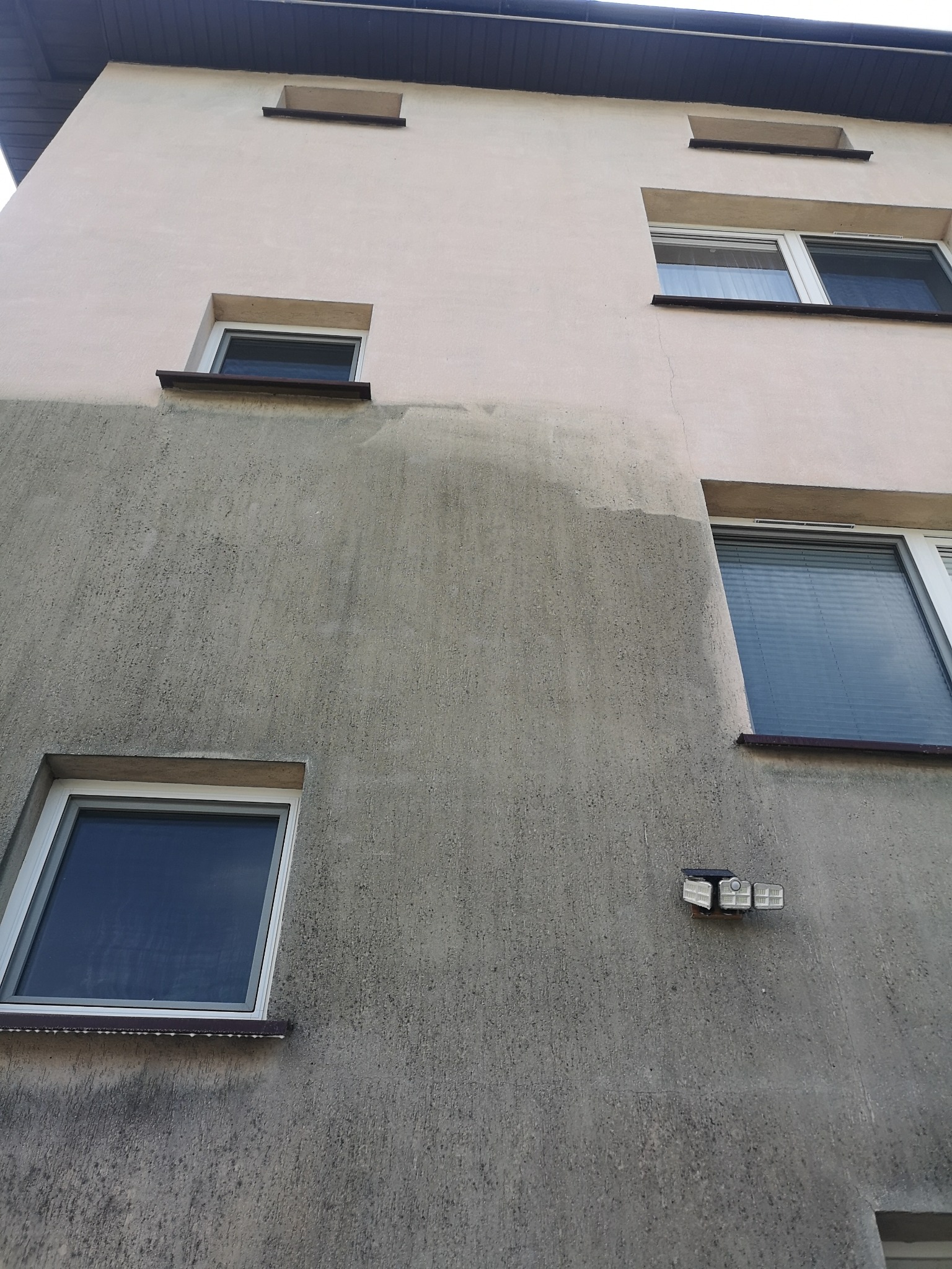 Fragment elewacji budynku z widocznym obszarem po czyszczeniu, kontrastującym z zabrudzoną częścią ściany, okna z brązowymi parapetami i lampa solarna na ścianie.