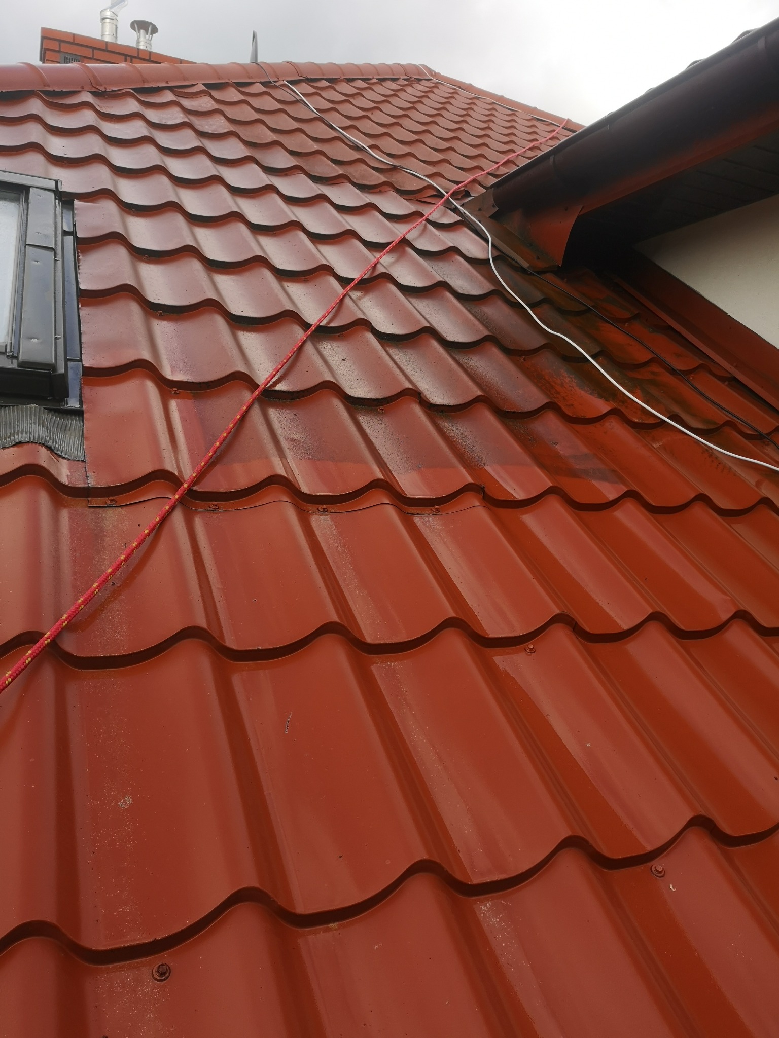 Czerwona blachodachówka, częściowo umyta, z widocznymi śladami zabrudzeń i liną zabezpieczającą biegnącą po skosie, okno dachowe po lewej stronie.