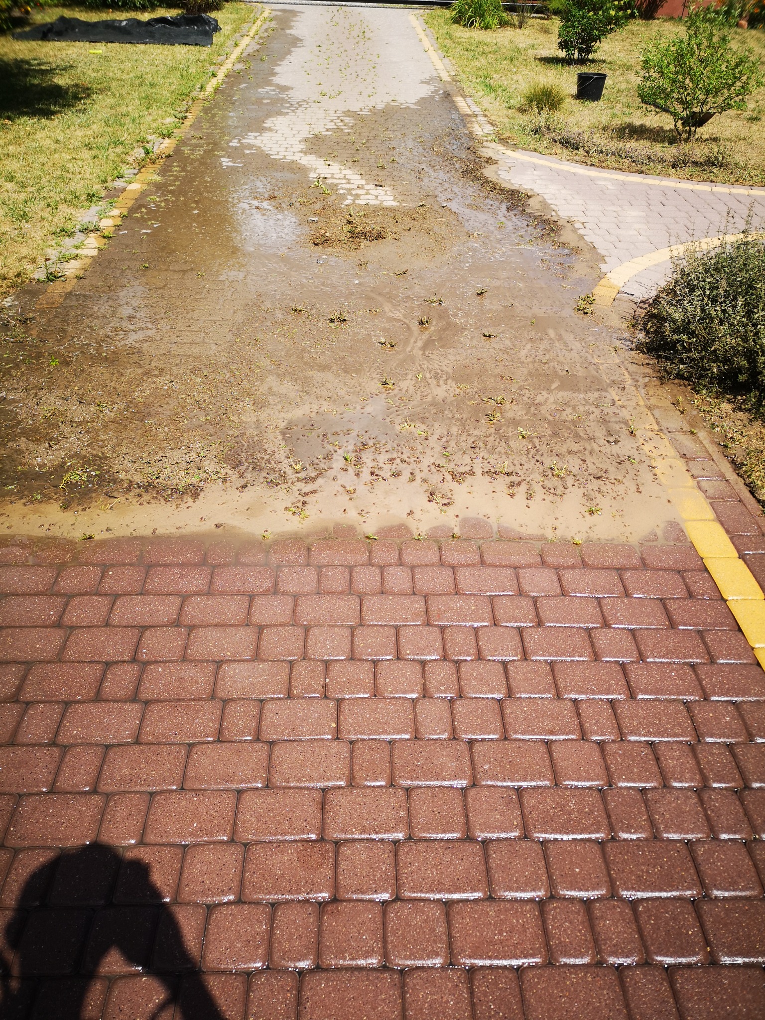 Częściowo umyta kostka brukowa z resztkami brudu i roślin, widoczny kontrast między czystą i brudną powierzchnią, cień osoby w dolnym rogu.