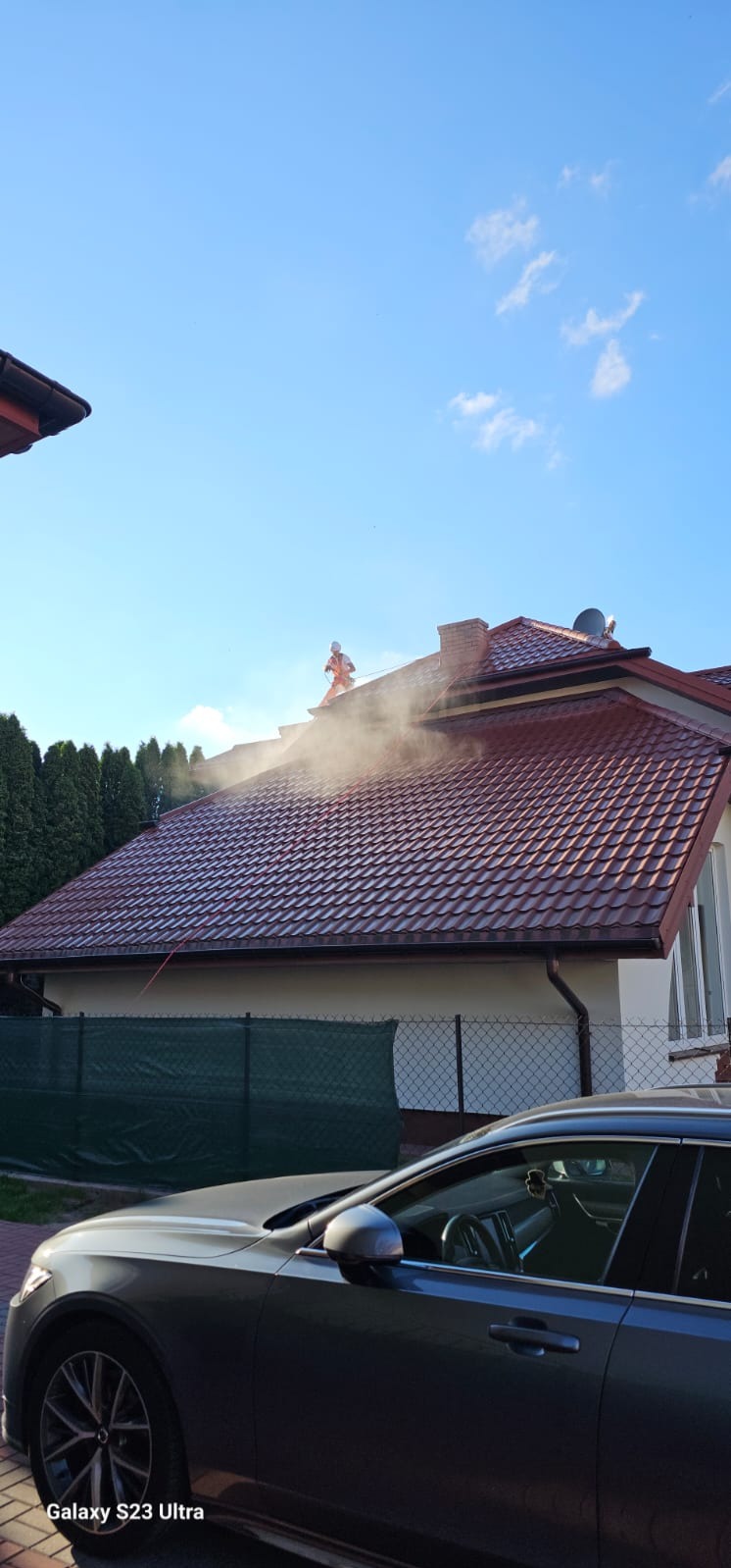 Pracownik w pomarańczowym kombinezonie czyści dach z czerwonej dachówki pod ciśnieniem, widoczna para wodna, w tle zielone tuje i fragment zaparkowanego szarego samochodu.