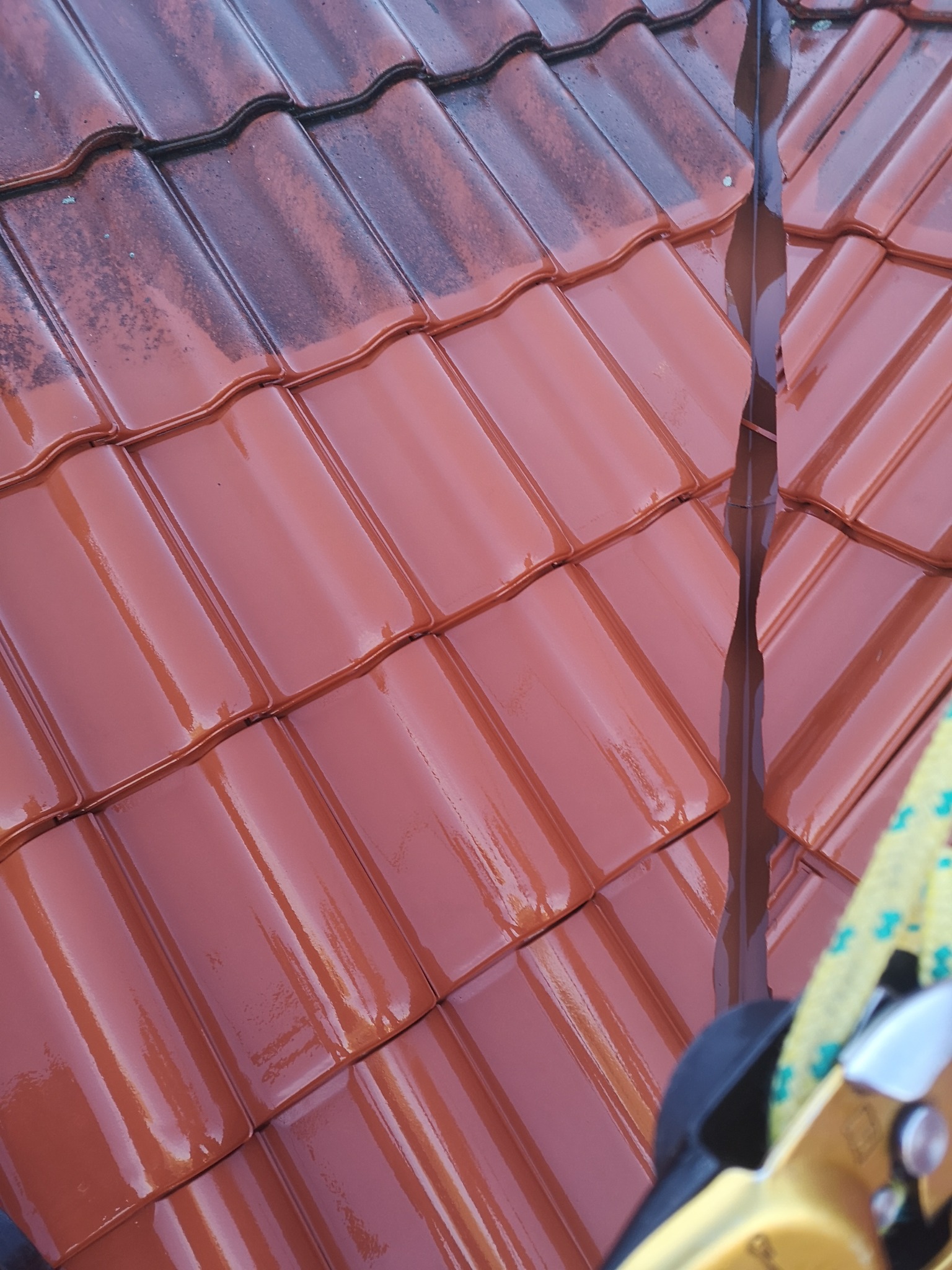 Czerwona dachówka ceramiczna, częściowo umyta, z widocznym zaciekami wody i fragmentem sprzętu asekuracyjnego, z bliska.