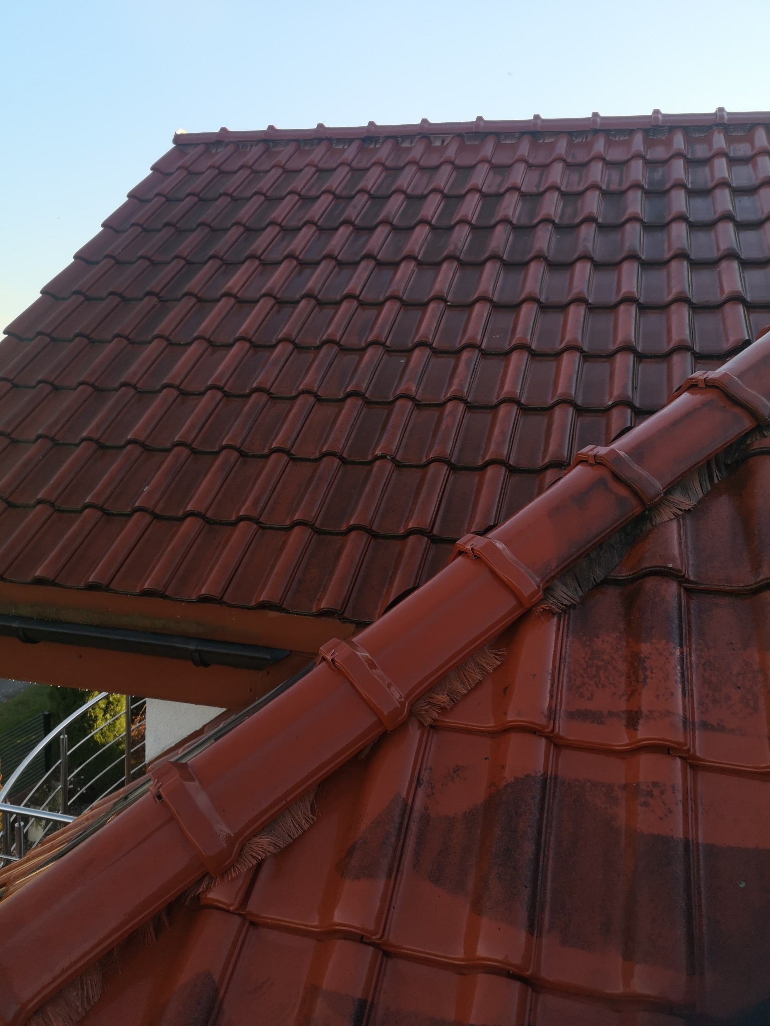 Czerwona dachówka ceramiczna, częściowo umyta, z widocznymi ciemniejszymi plamami zabrudzeń, obok rynna z zamontowanym zabezpieczeniem przed liśćmi.