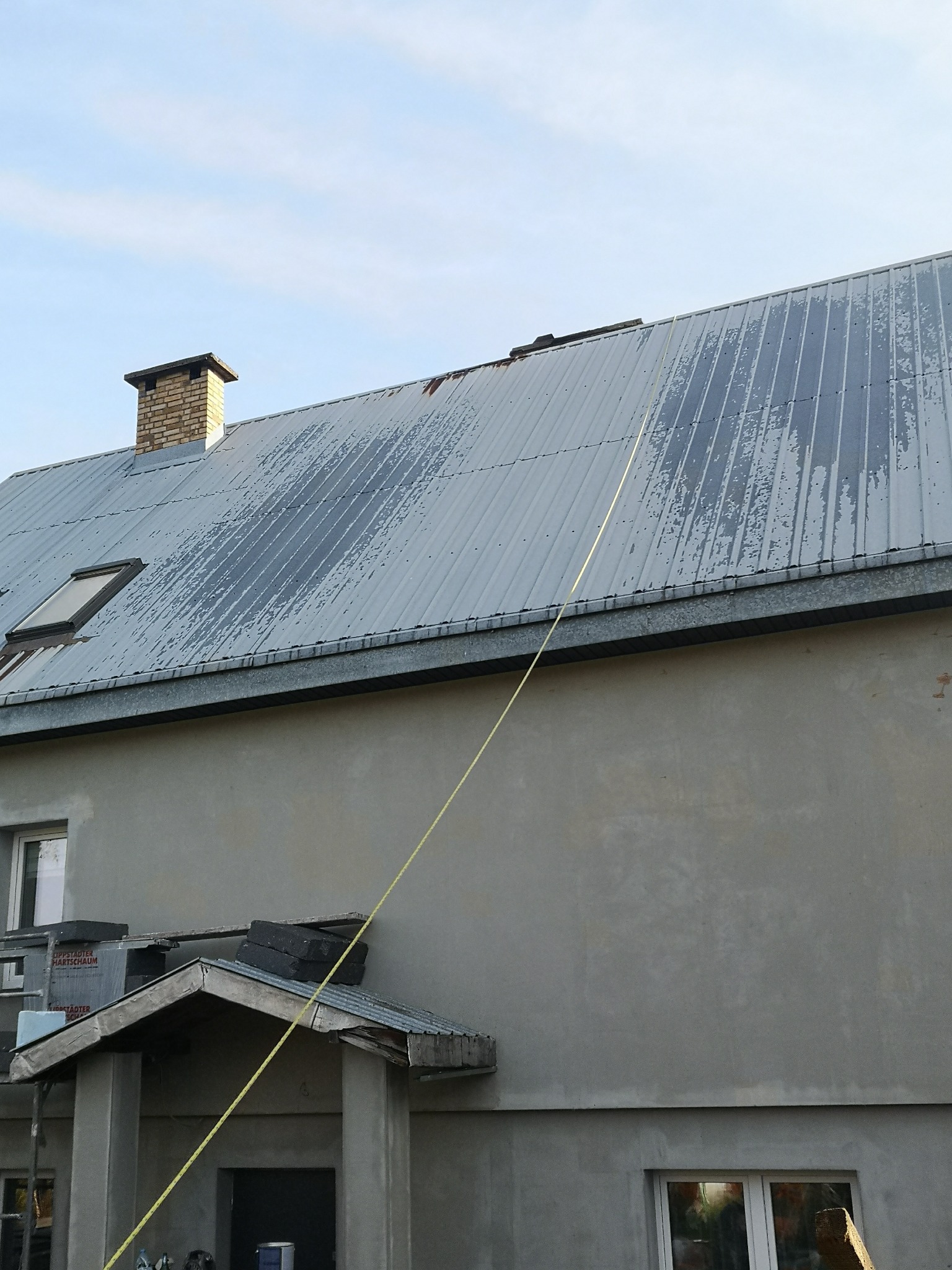 Szary, blaszany dach z widocznymi śladami korozji i zabrudzeń, z kominem i oknem dachowym, na tle jasnego nieba i szarej ściany budynku z zadaszeniem wejściowym, z żółtą liną biegnącą po dachu.