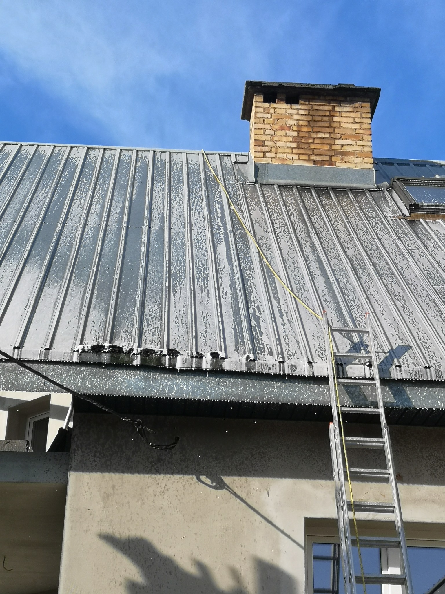 Metalowy dach po umyciu, z widocznymi smugami wody, drabina oparta o budynek, ceglany komin na tle błękitnego nieba.