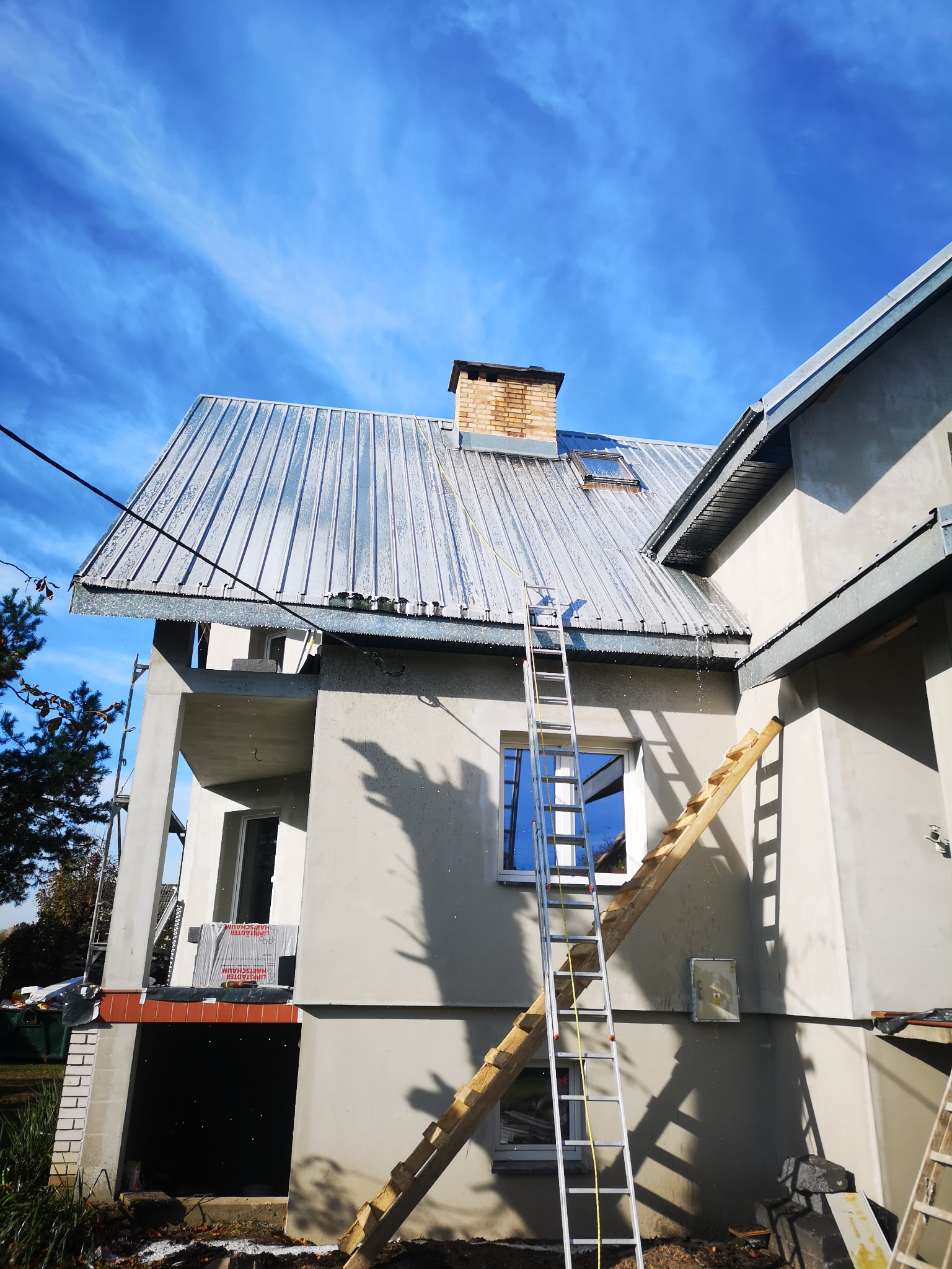 Dwupiętrowy dom w trakcie budowy z metalowym dachem, na którym widoczne są ślady wody. Do ściany oparta drabina, a w tle błękitne niebo z delikatnymi chmurami.