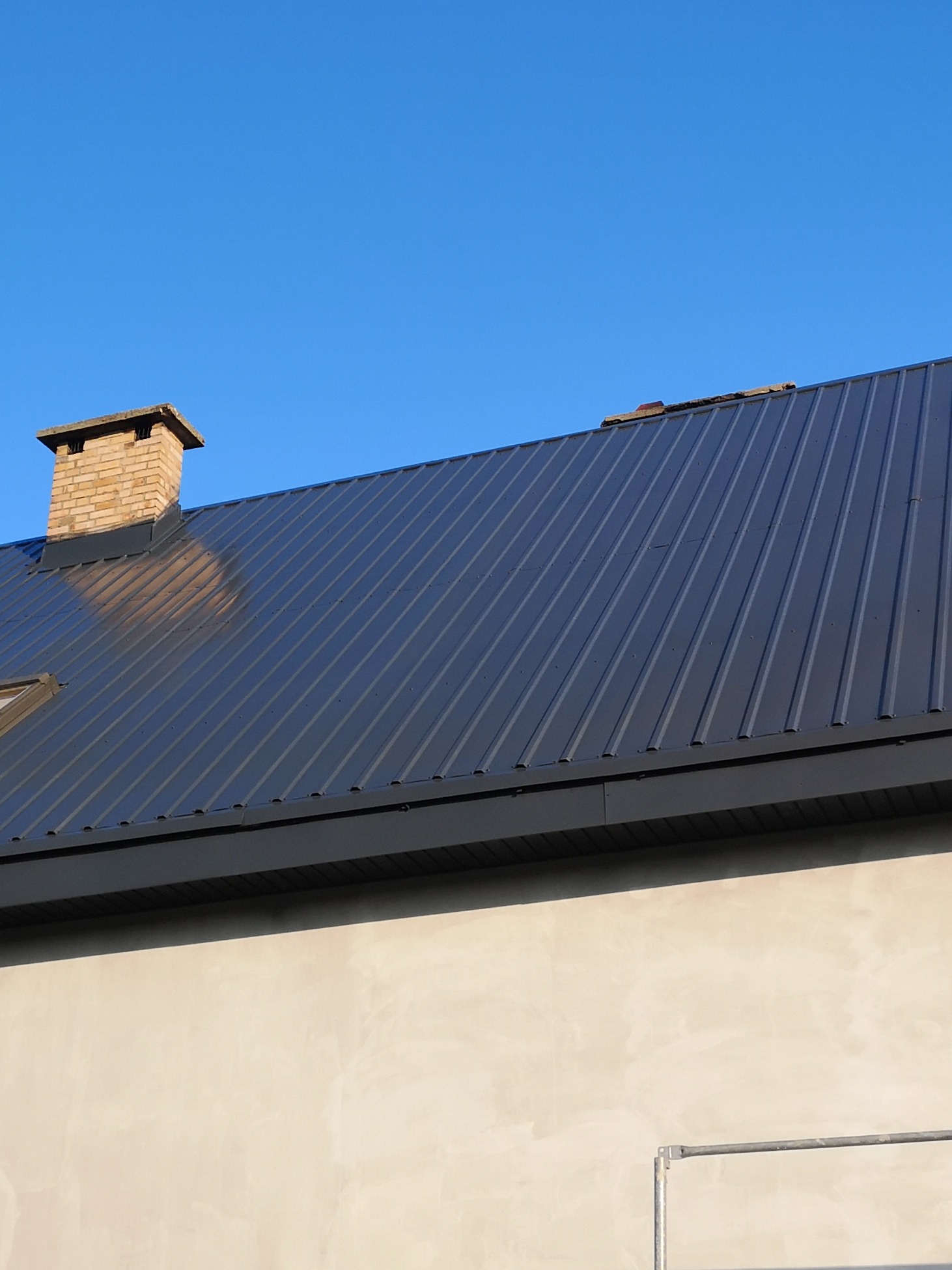 Ciemnoszary dach z blachy trapezowej, ceglany komin z daszkiem, fragment otynkowanej ściany budynku i fragment rusztowania na tle błękitnego nieba.