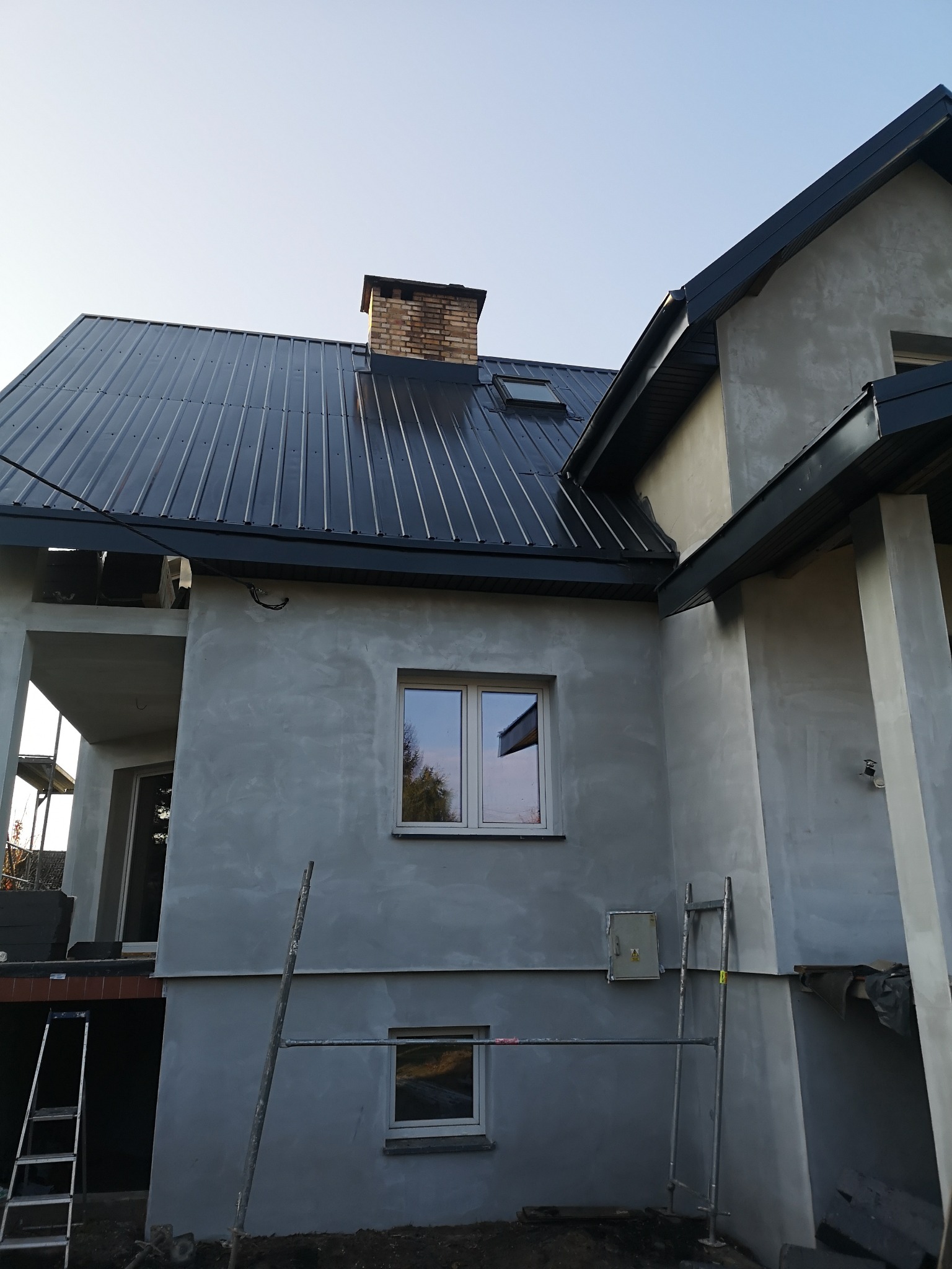 Dom w trakcie budowy lub remontu, z nowym, błyszczącym, ciemnym dachem z blachy trapezowej, widoczne rusztowania i surowa elewacja w kolorze szarym.