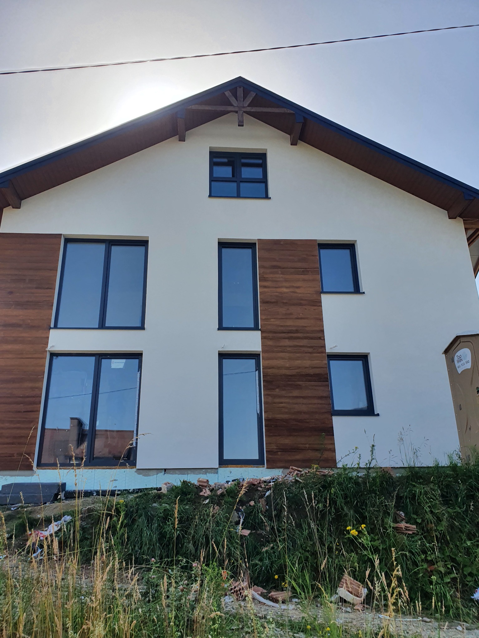 Dwupiętrowy dom z białą elewacją, drewnianymi panelami bocznymi i ciemnymi ramami okiennymi, widziany z dołu w trakcie prac wykończeniowych, z widocznymi pozostałościami materiałów budowlanych...