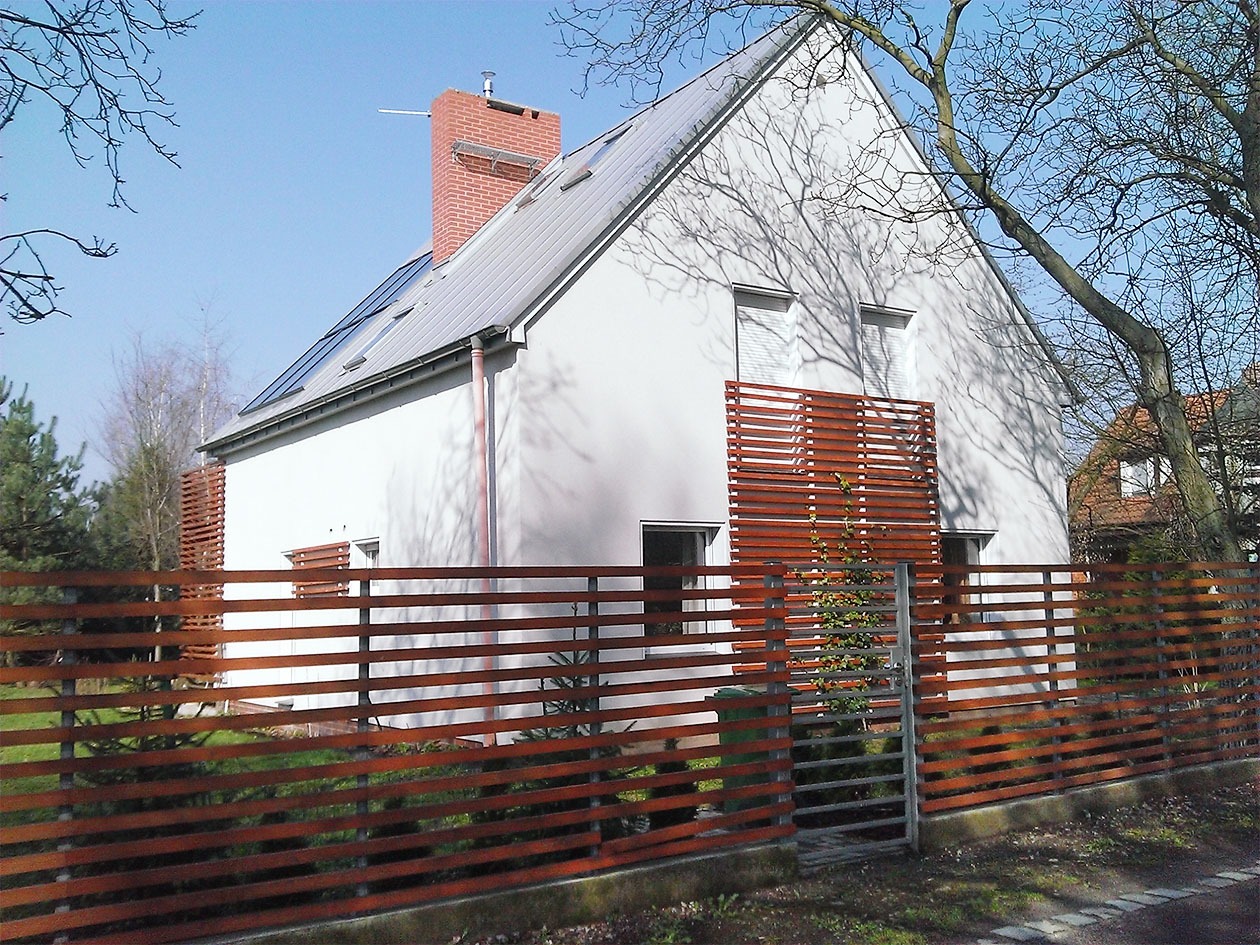 Nowoczesny dom jednorodzinny z białymi ścianami, szarym dachem i ceglanym kominem, otoczony drewnianym płotem z poziomymi deskami i elementami przesłaniającymi okna.