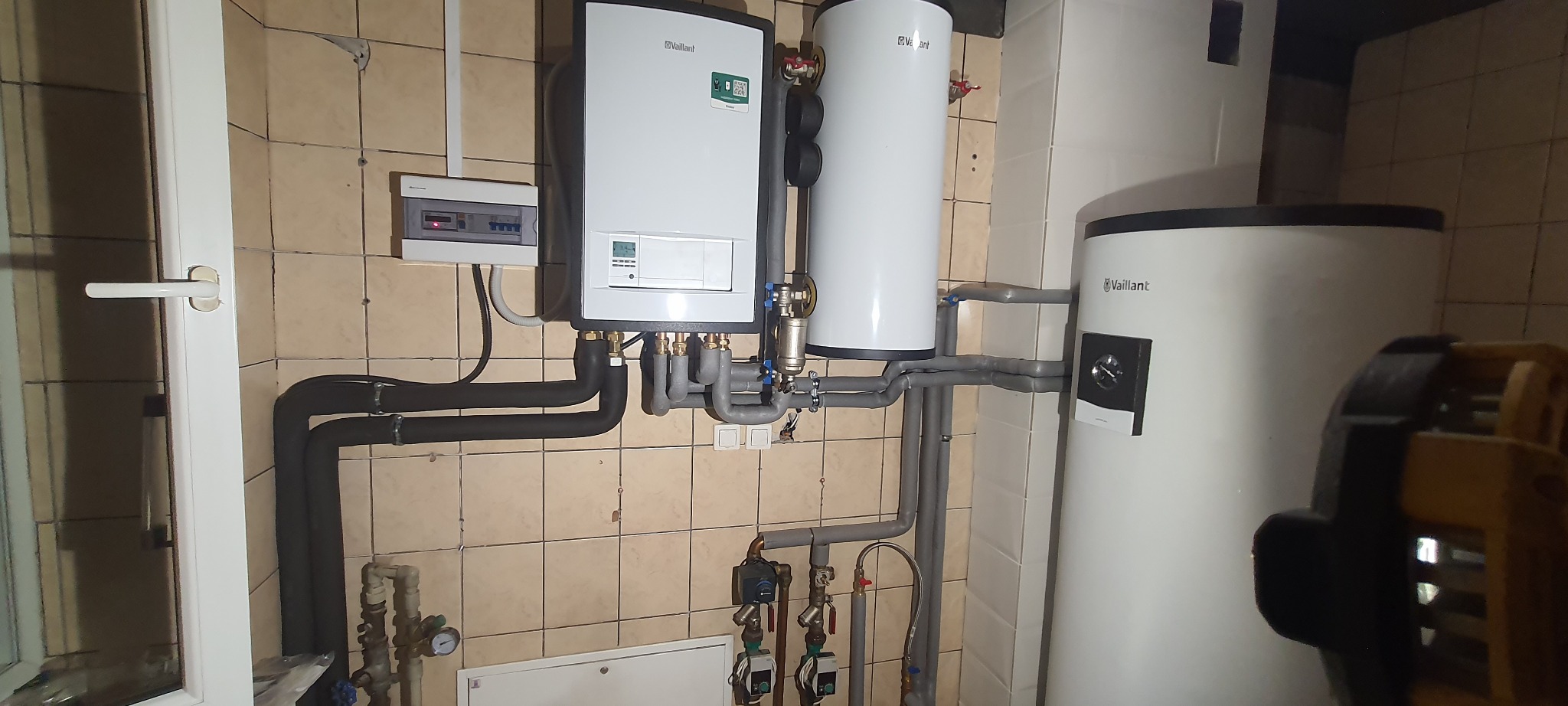 Kotłownia z piecem gazowym Vaillant, zasobnikiem ciepłej wody i rozdzielnicą elektryczną na ścianie z beżowych płytek.