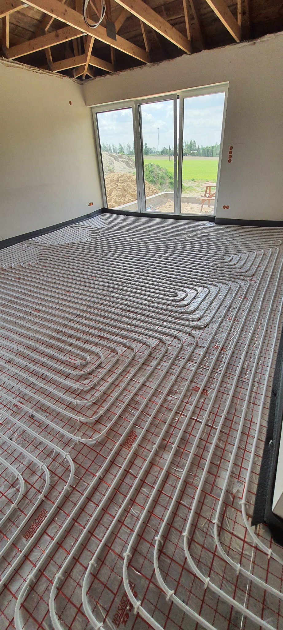 Białe rurki ogrzewania podłogowego ułożone w pętle na czerwonej siatce w surowym wnętrzu z widokiem na zewnątrz przez okno.
