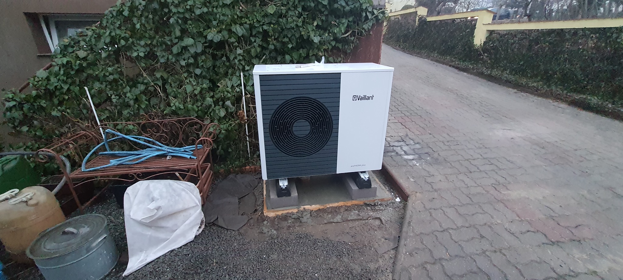 Biała pompa ciepła Vaillant AROTHERM plus, zamontowana na betonowych bloczkach, ustawiona na posesji obok chodnika z kostki brukowej, z widocznymi elementami ogrodu w tle.