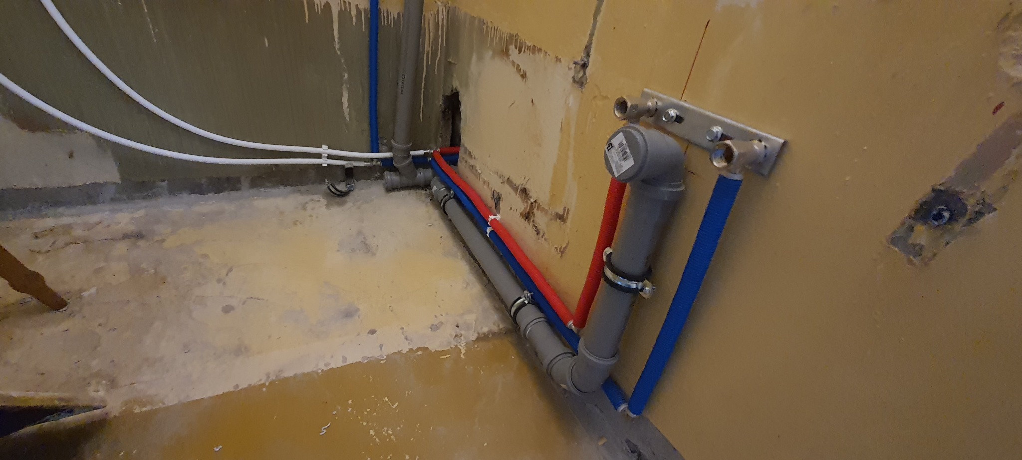 Fragment instalacji hydraulicznej w trakcie montażu, widoczne rury PEX w kolorze czerwonym i niebieskim, szare rury PCV, metalowa listwa mocująca z przyłączami, ściana w trakcie remontu.