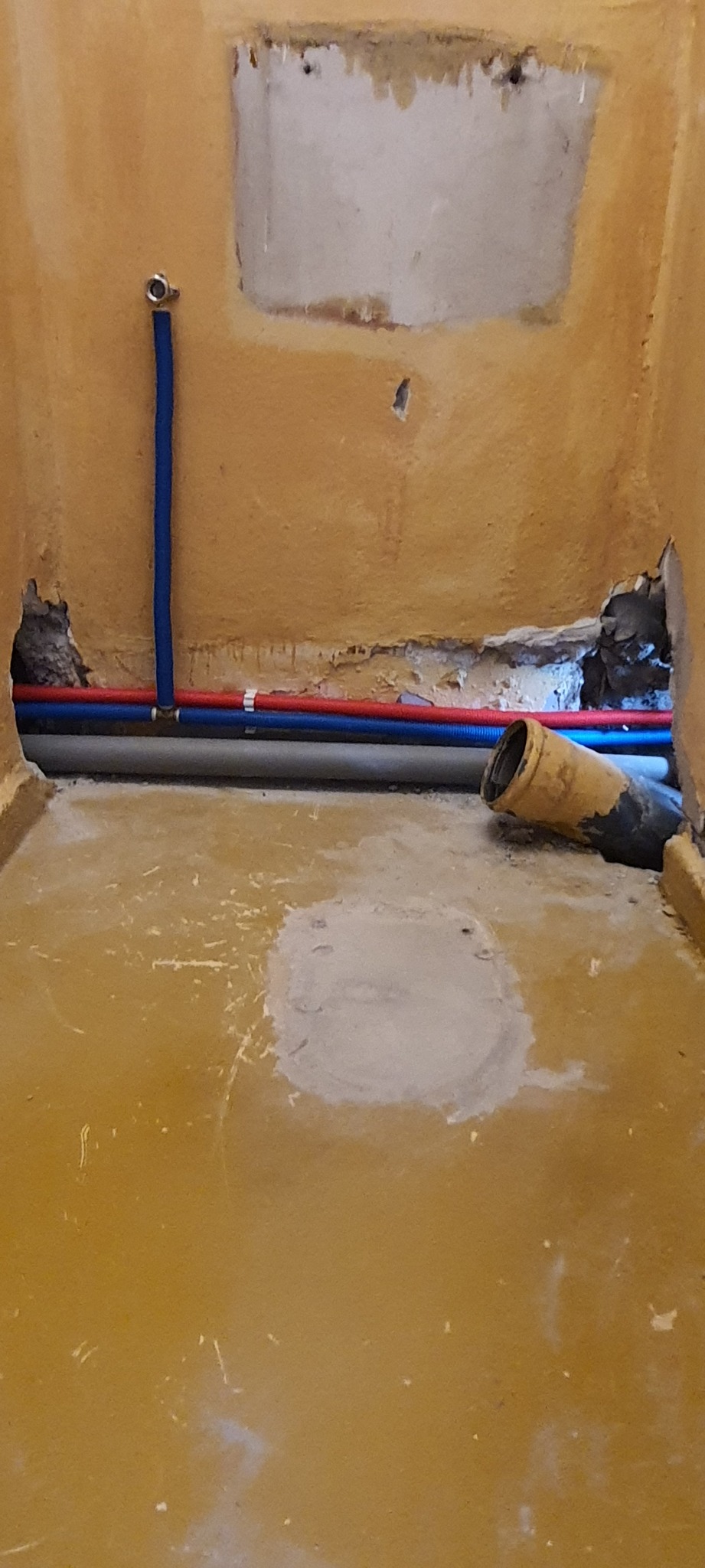 Przygotowanie do instalacji hydraulicznej: widoczne rury PEX w kolorach niebieskim i czerwonym, szara rura kanalizacyjna z żółtymi elementami, oraz fragment ściany z widocznymi uszkodzeniami...