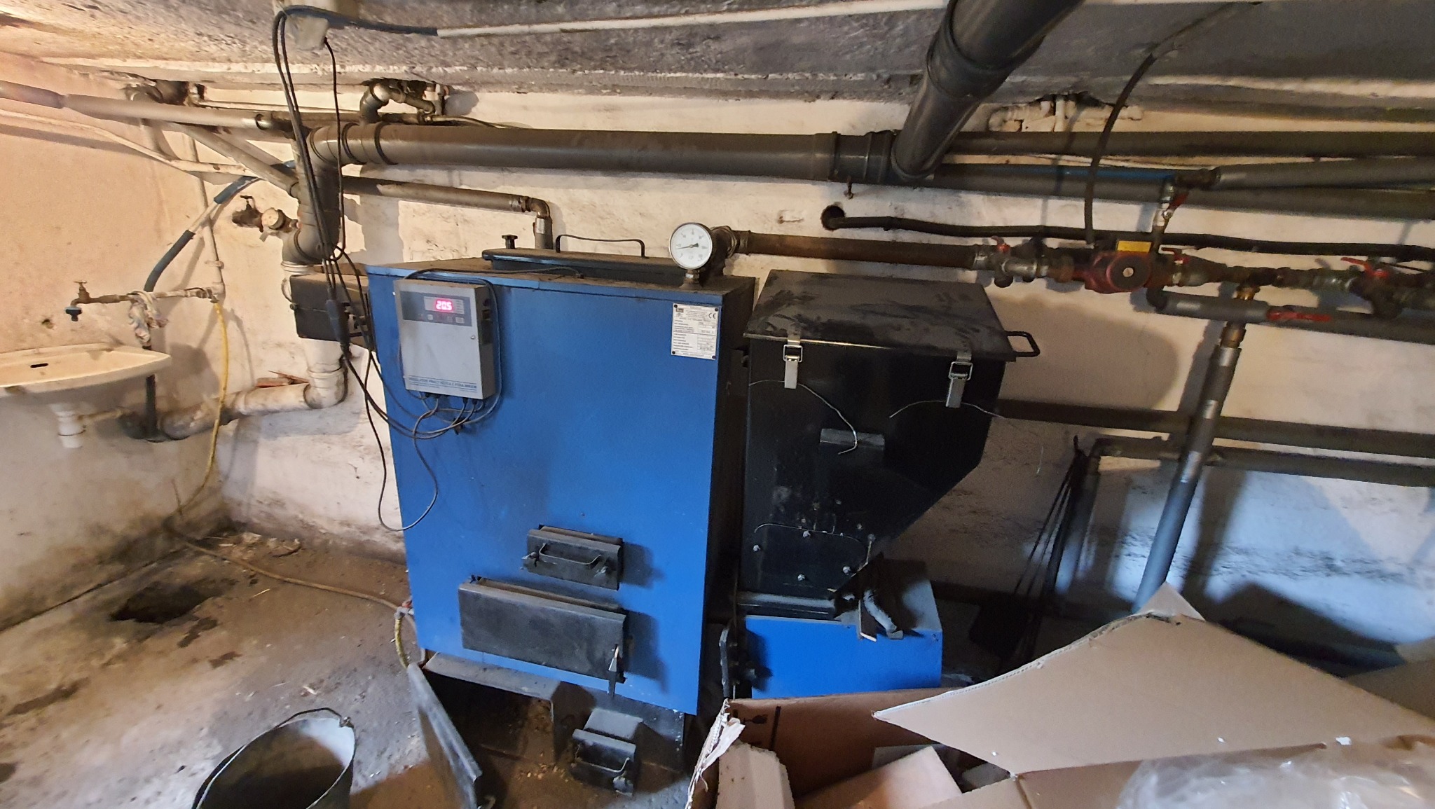 Kotłownia z kotłem na paliwo stałe, widoczne rury instalacji CO, manometr, sterownik i zlew w piwnicy.