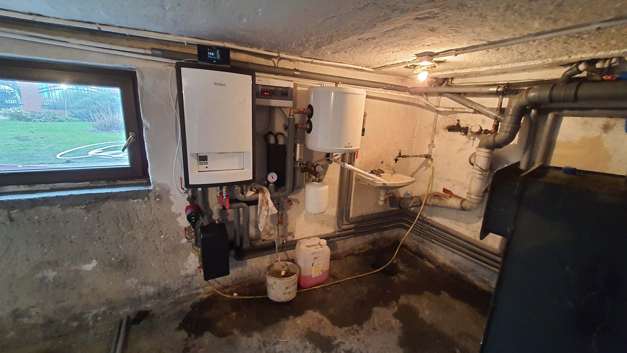 Piwnica z zainstalowanym, białym piecem gazowym Vaillant, bojlerem i licznymi rurami. Widoczne okno z widokiem na ogród. Na podłodze wiadro z płynem.