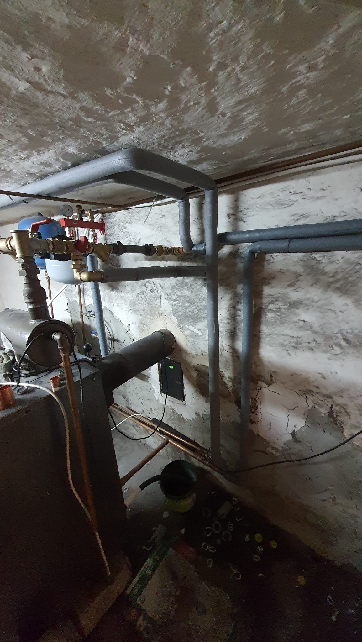 Kotłownia w piwnicy: instalacja centralnego ogrzewania z rurami izolowanymi termicznie, widoczne elementy kotła, armatura i fragment ściany z odpadającym tynkiem.