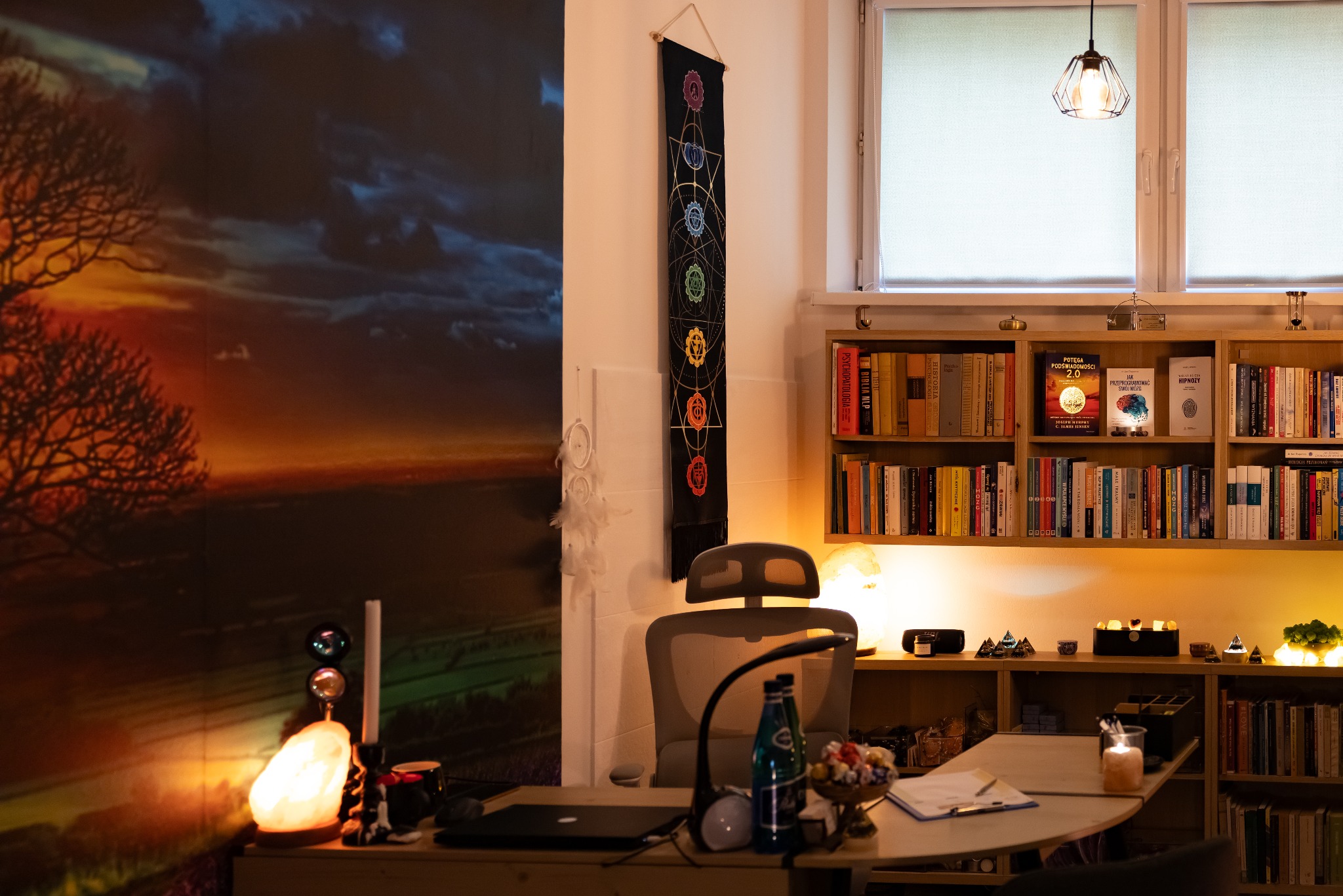 Gabinet: biurko z laptopem, lampka, dekoracje, książki na półkach, plakat z czakrami. Przytulne, profesjonalne wnętrze do sesji.