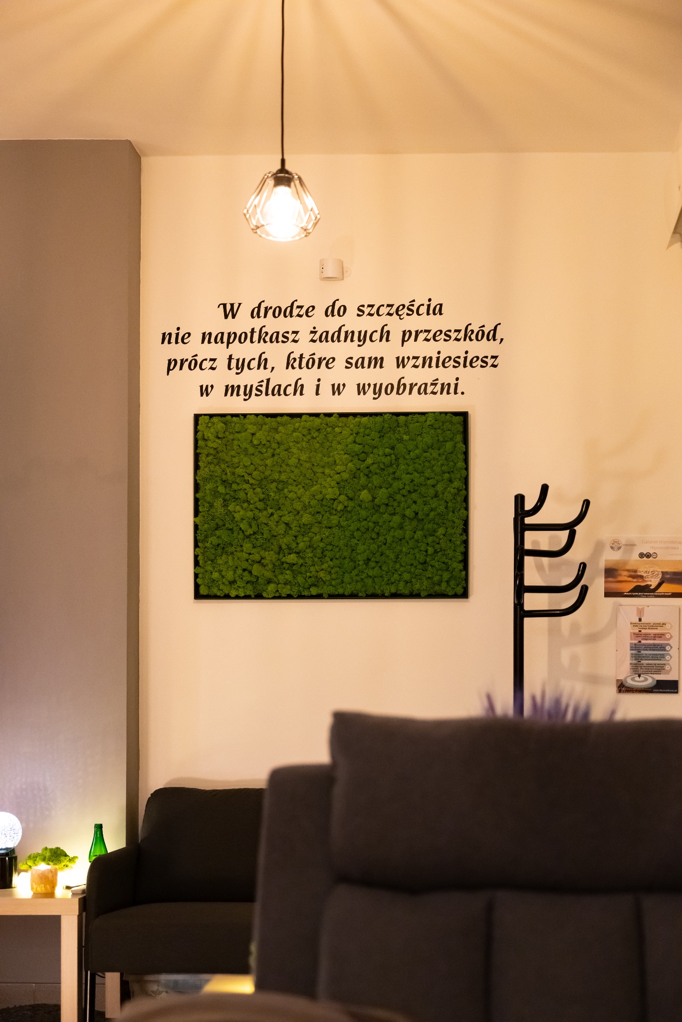 Wnętrze gabinetu hipnoterapii z sentencją na ścianie, panelem z mchu, lampą i elementami wyposażenia, tworzące spokojną atmosferę. Przytulny wystrój.