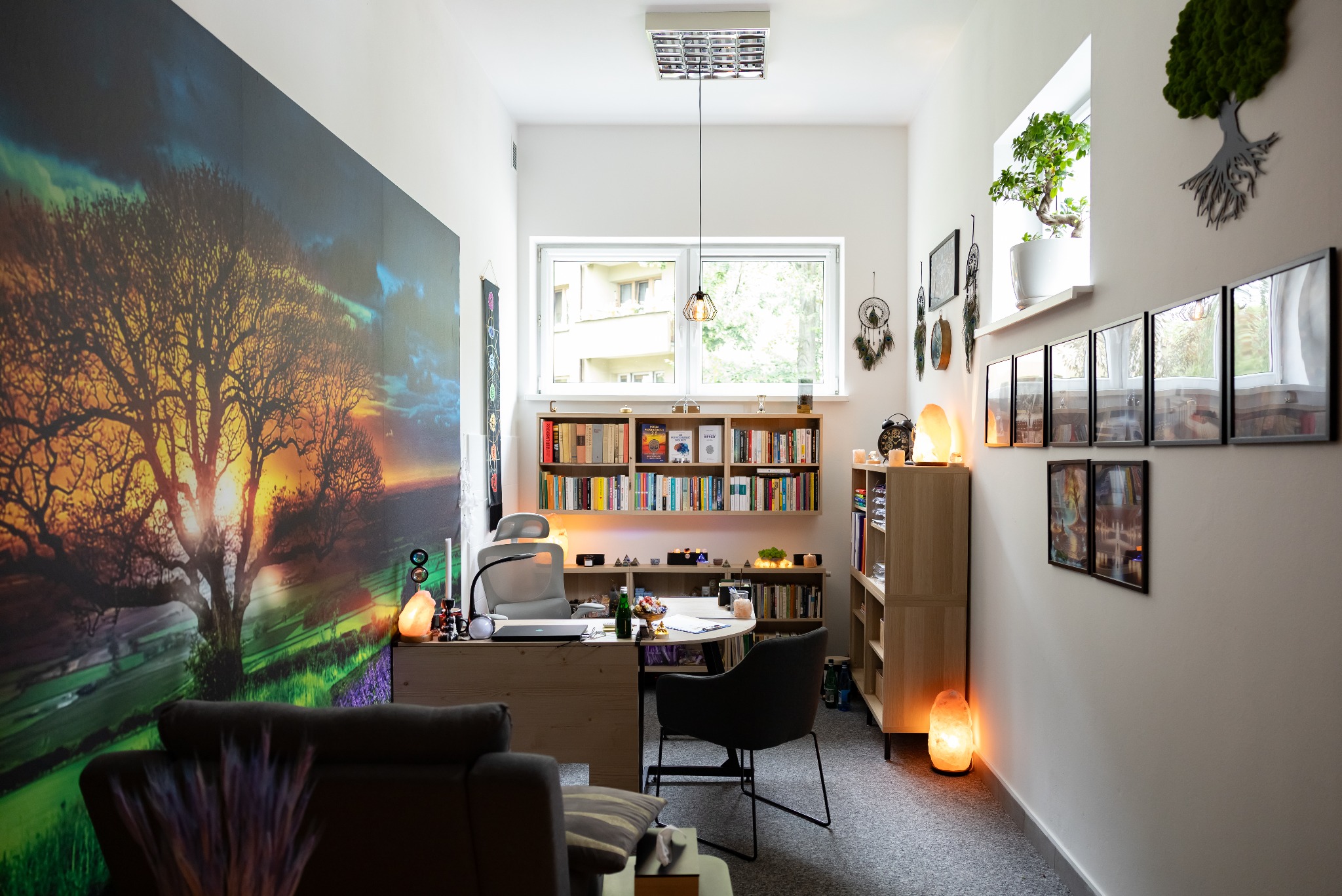 Jasne wnętrze gabinetu z biurkiem, regałami i dekoracjami, w tym solne lampy i obraz drzewa na ścianie. Przytulna przestrzeń do sesji relaksacyjnych.
