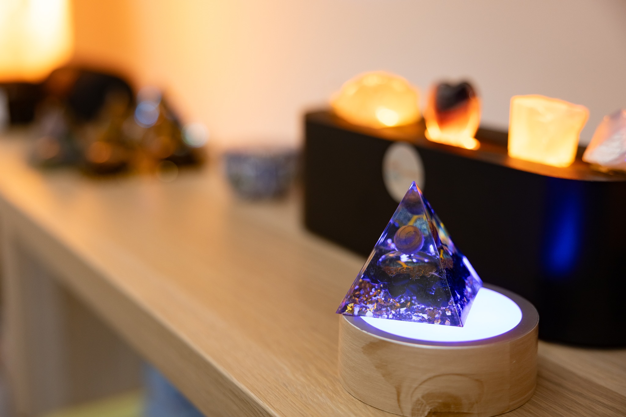 Szklana piramida z zatopionymi elementami na podświetlanej podstawce, obok podświetlane kamienie w czarnej obudowie. Ciepłe, rozproszone światło w tle.