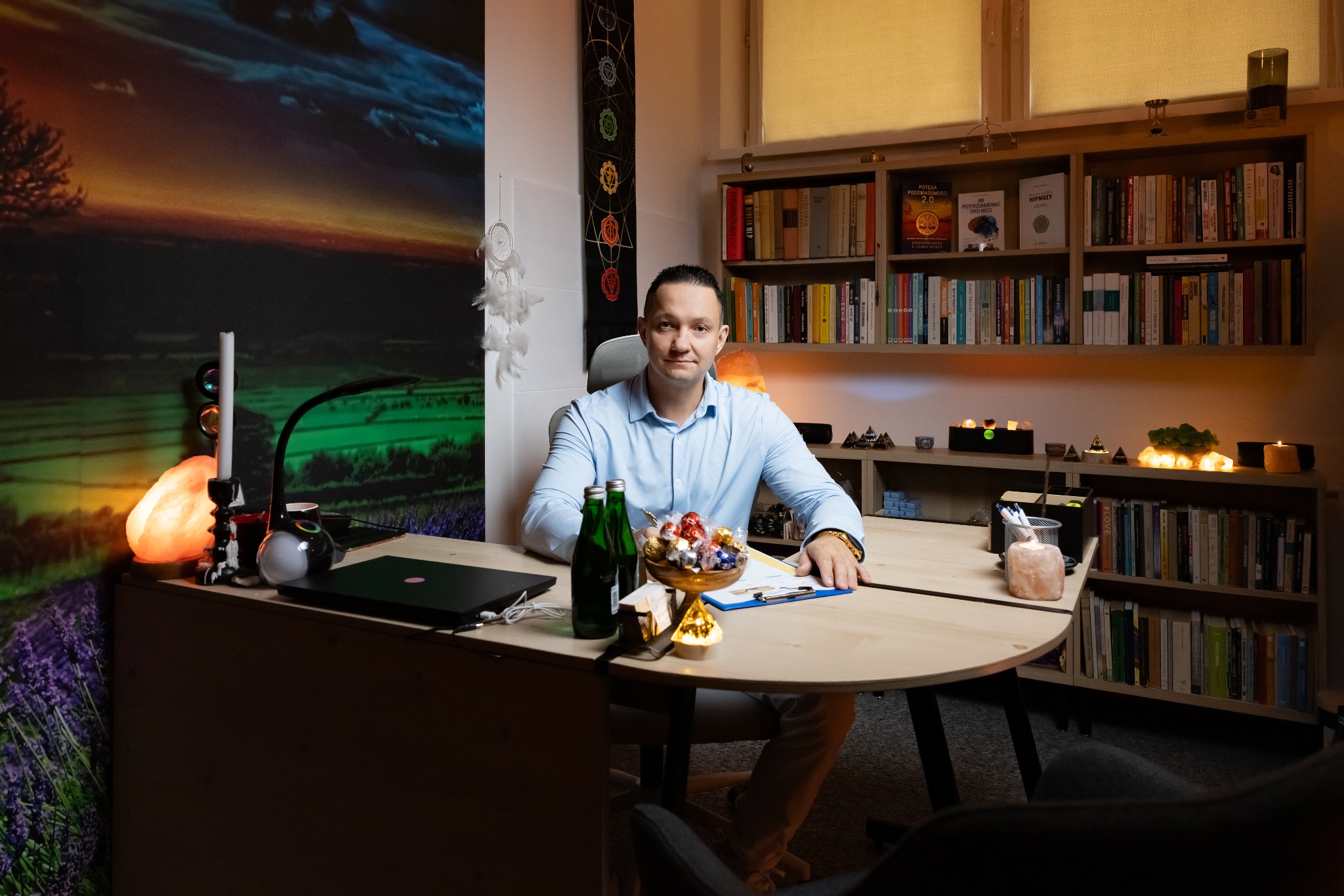 Mężczyzna w błękitnej koszuli siedzi za biurkiem w gabinecie z książkami, lampami solnymi i dekoracjami, przygotowany do sesji.