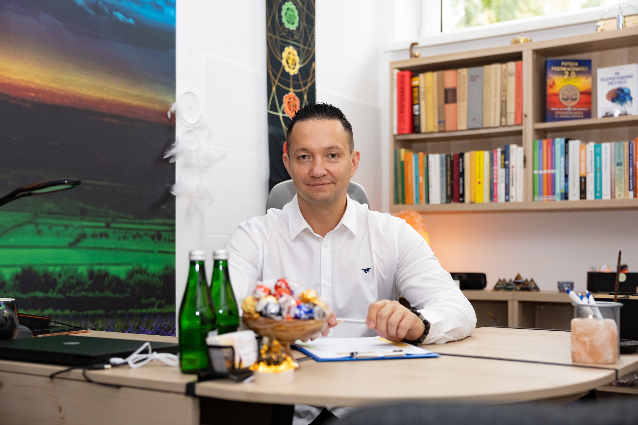 Mężczyzna w białej koszuli siedzi za biurkiem w gabinecie. Na biurku słodycze, woda, notatnik. W tle regał z książkami i dekoracje związane z duchowością.