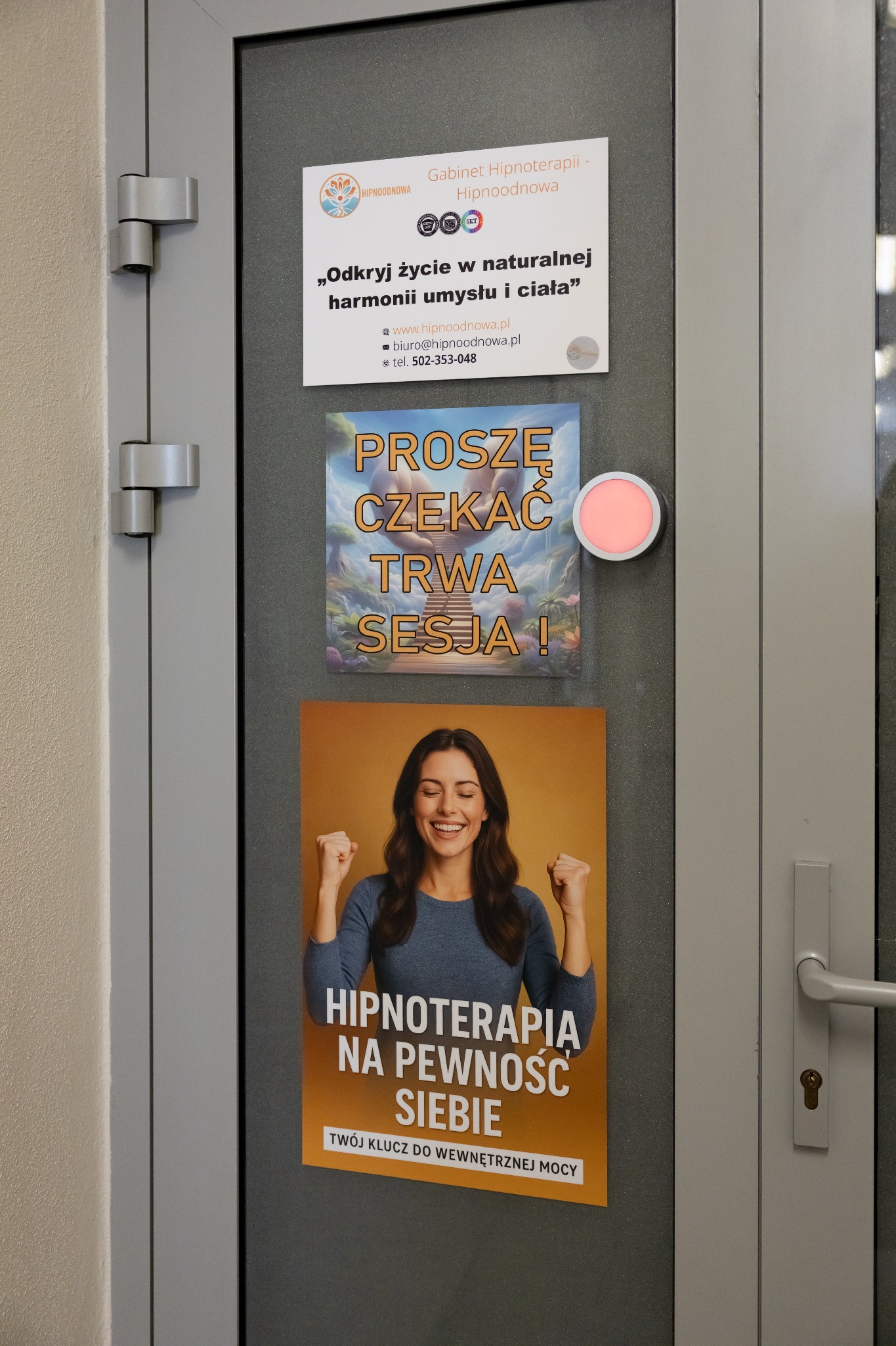 Drzwi gabinetu z ogłoszeniami: oferta hipnoterapii, informacja o trwającej sesji i dane kontaktowe. Plakaty w stonowanych kolorach. Widoczny włącznik światła.