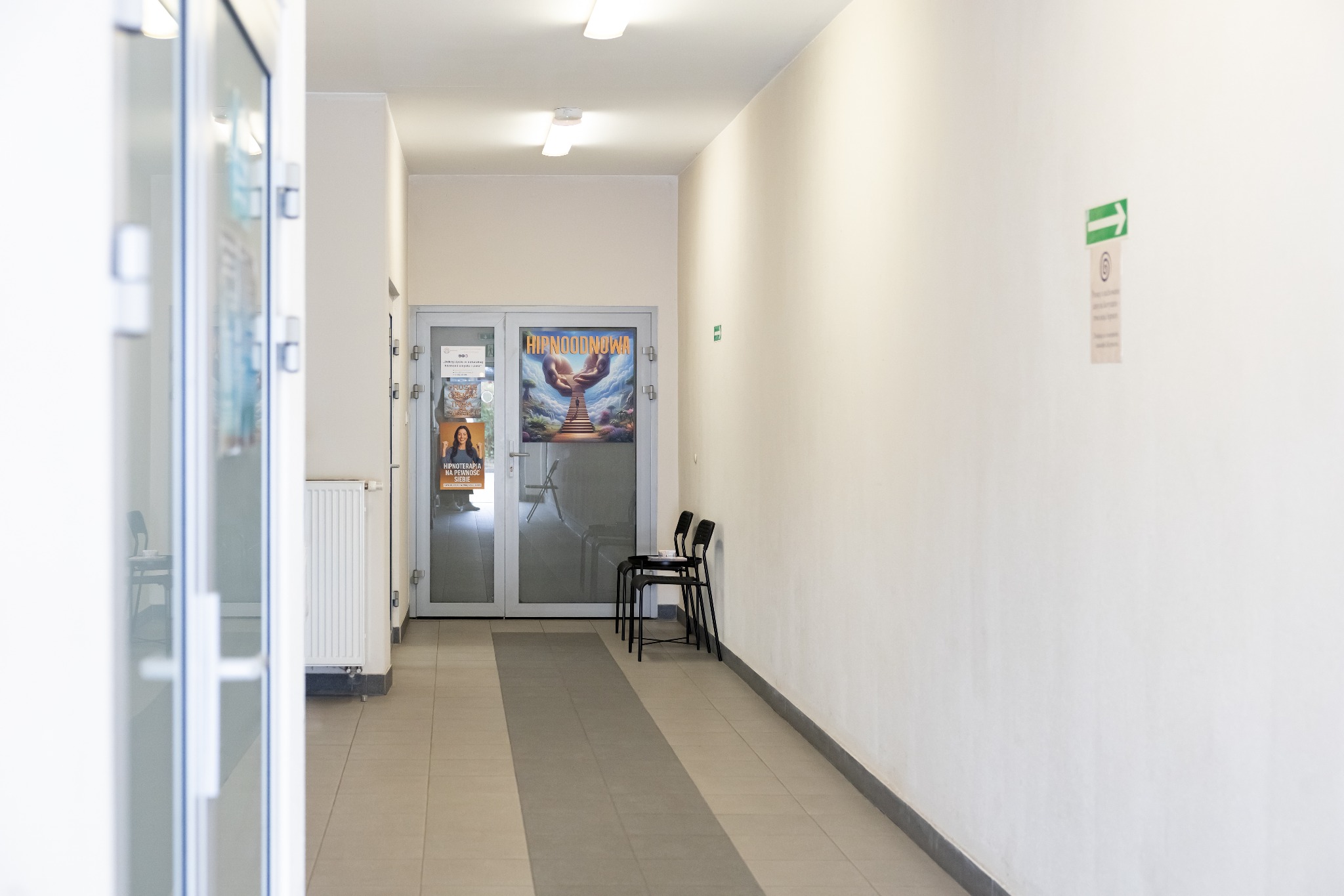 Jasne wnętrze poczekalni z plakatem na szklanych drzwiach i dwoma krzesłami. Na ścianie zielona strzałka informacyjna.
