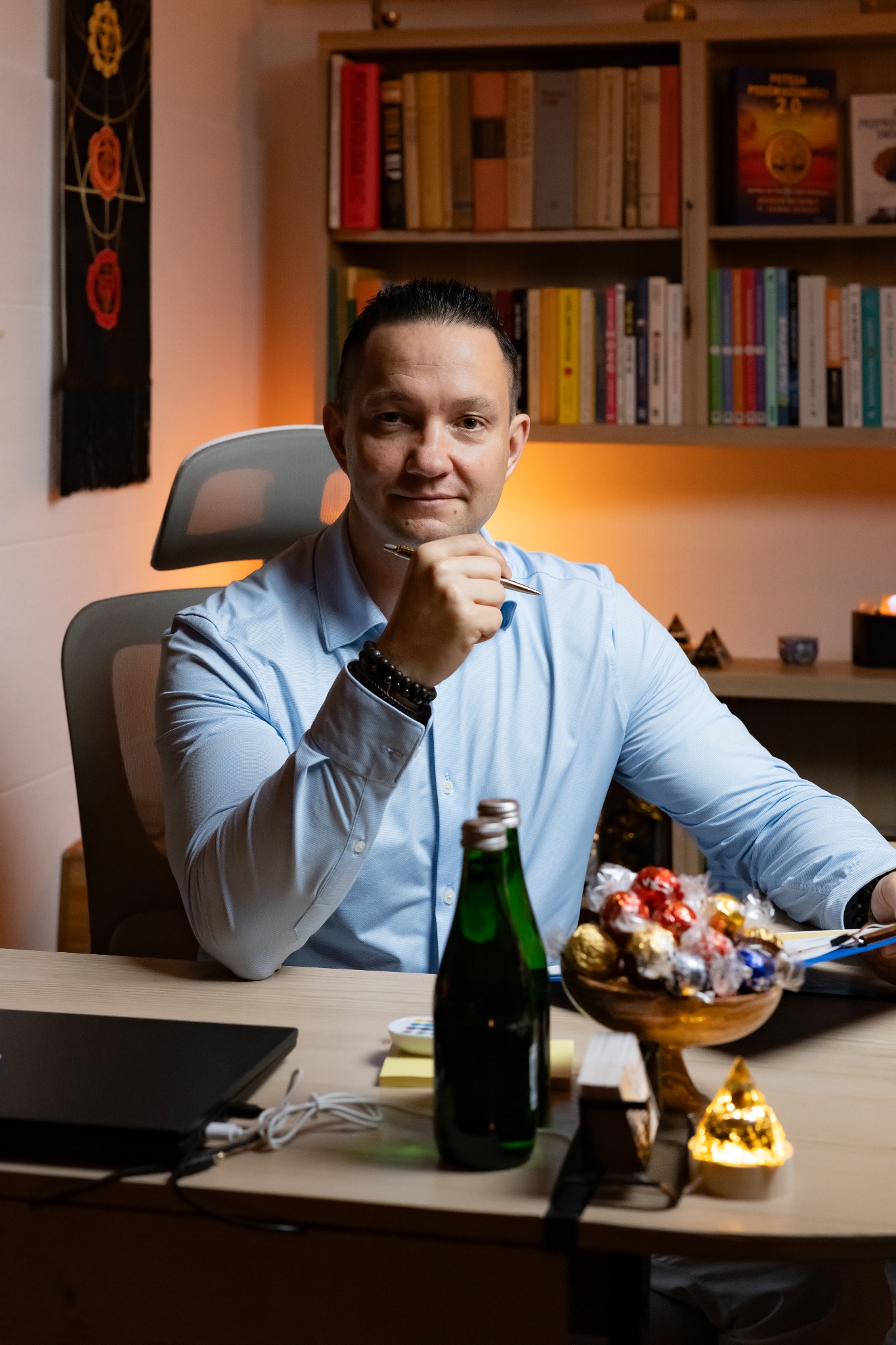 Portret mężczyzny w błękitnej koszuli siedzącego przy biurku z książkami w tle, trzymającego długopis przy brodzie, w gabinecie.