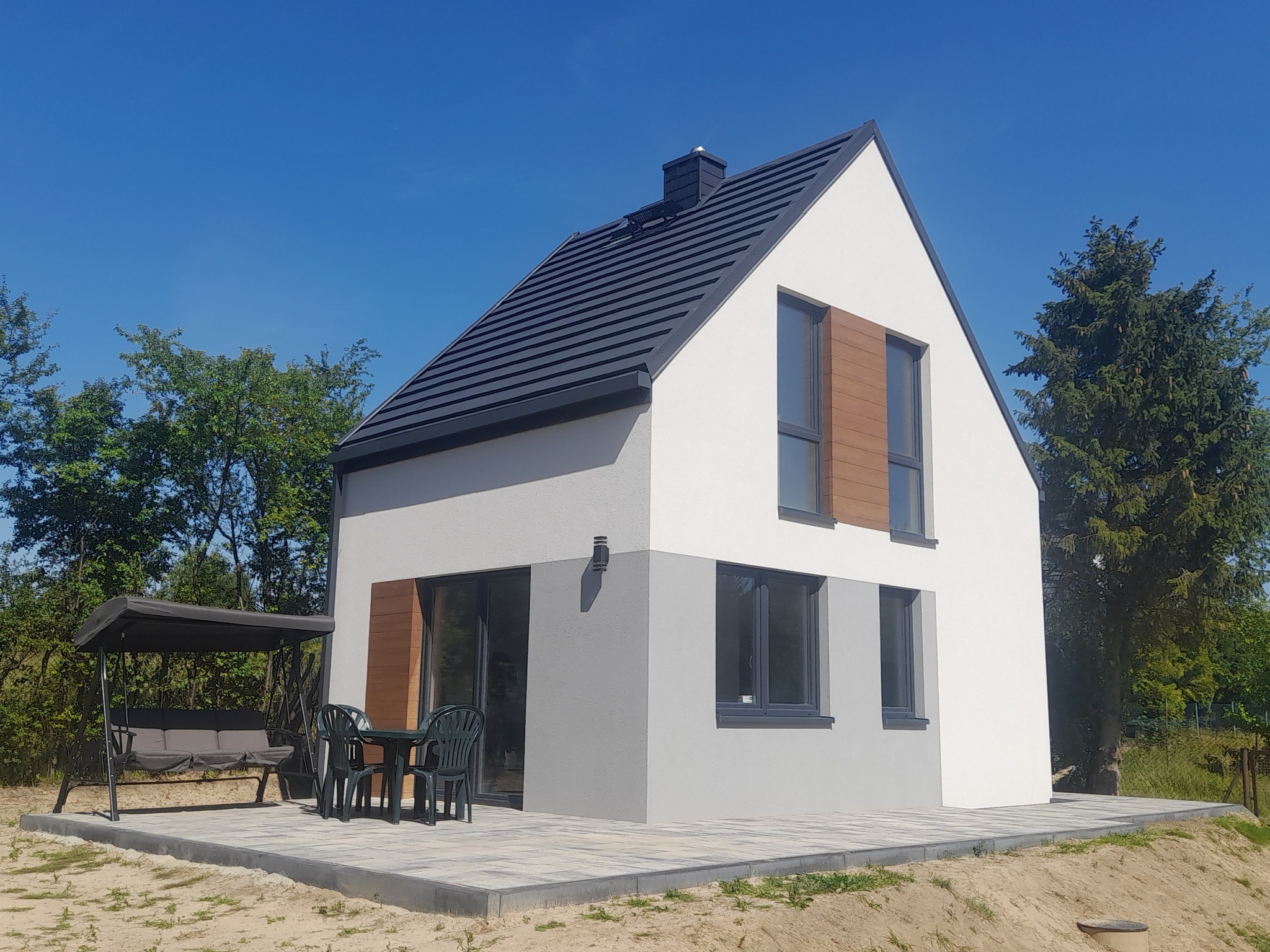 Nowoczesny dom jednorodzinny z dwukolorową elewacją (biało-szarą), ciemną dachówką i drewnopodobnymi żaluzjami, otoczony zielenią i zadaszoną huśtawką ogrodową na kostce brukowej.