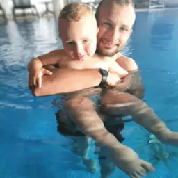 Uśmiechnięty mężczyzna z brodą trzymający małego chłopca w basenie z błękitną wodą i białymi kafelkami w tle, widoczny zegarek na ręce mężczyzny.